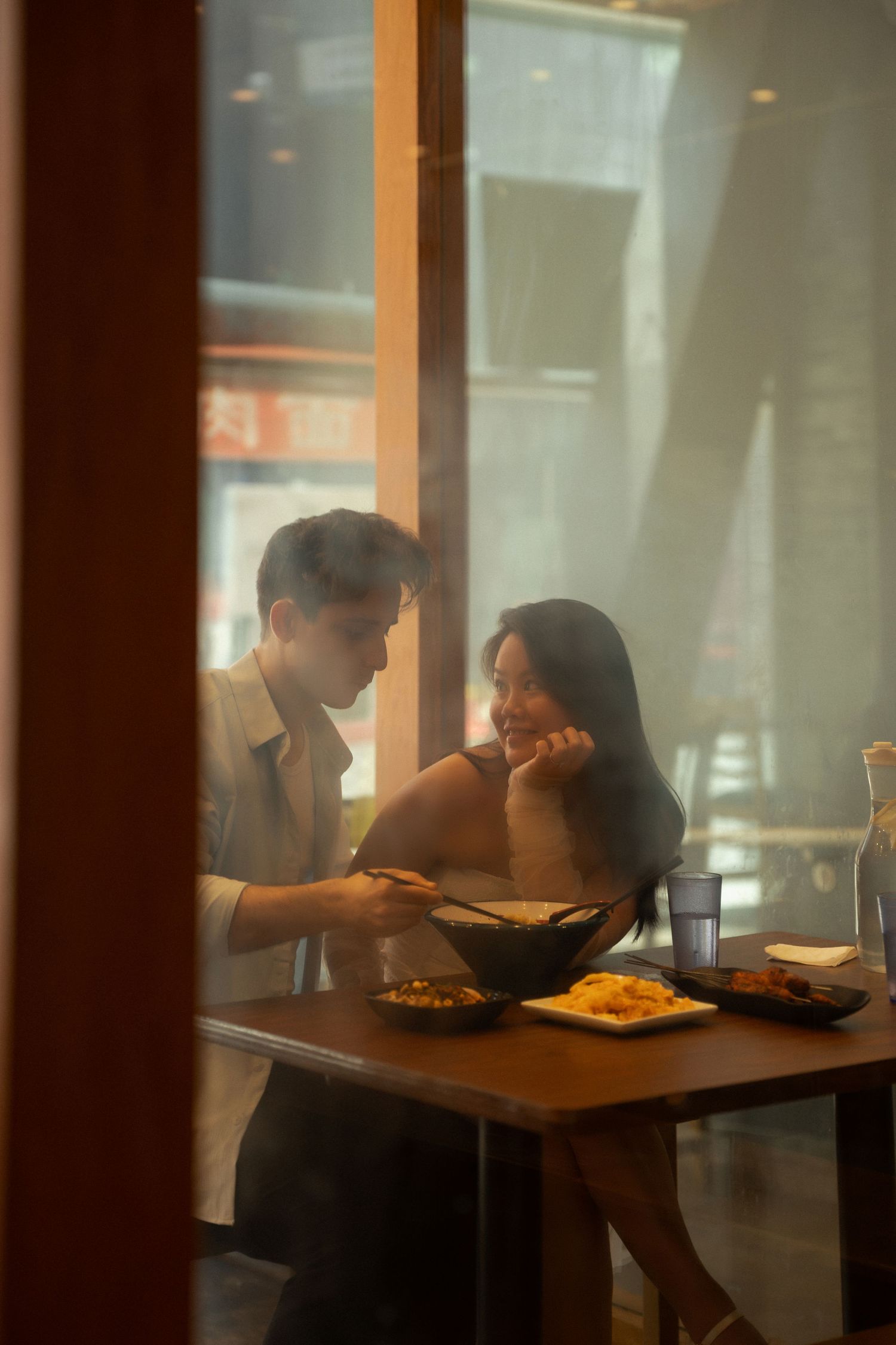 Couple sharing an intimate dinner date at a cozy restaurant, viewed through a window with warm ambient lighting.
