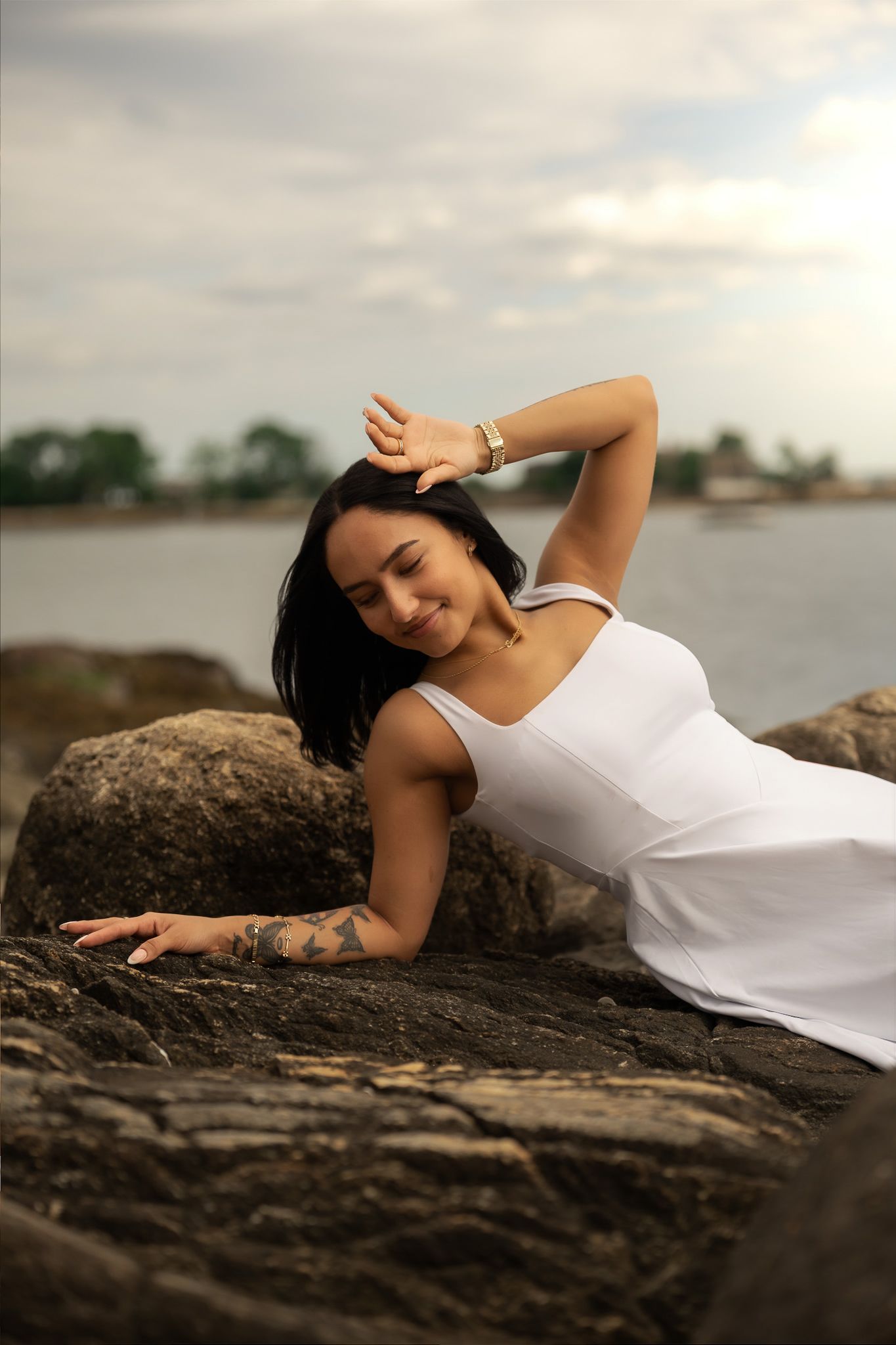Woman in white dress posing gracefully on rocky shore, smiling with hand raised, tattoo visible, ocean backdrop.