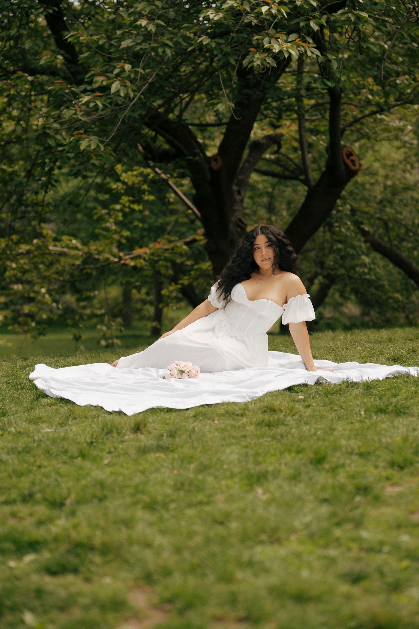 A bride in a flowing white off-shoulder gown sits gracefully on a white blanket in a lush green park.