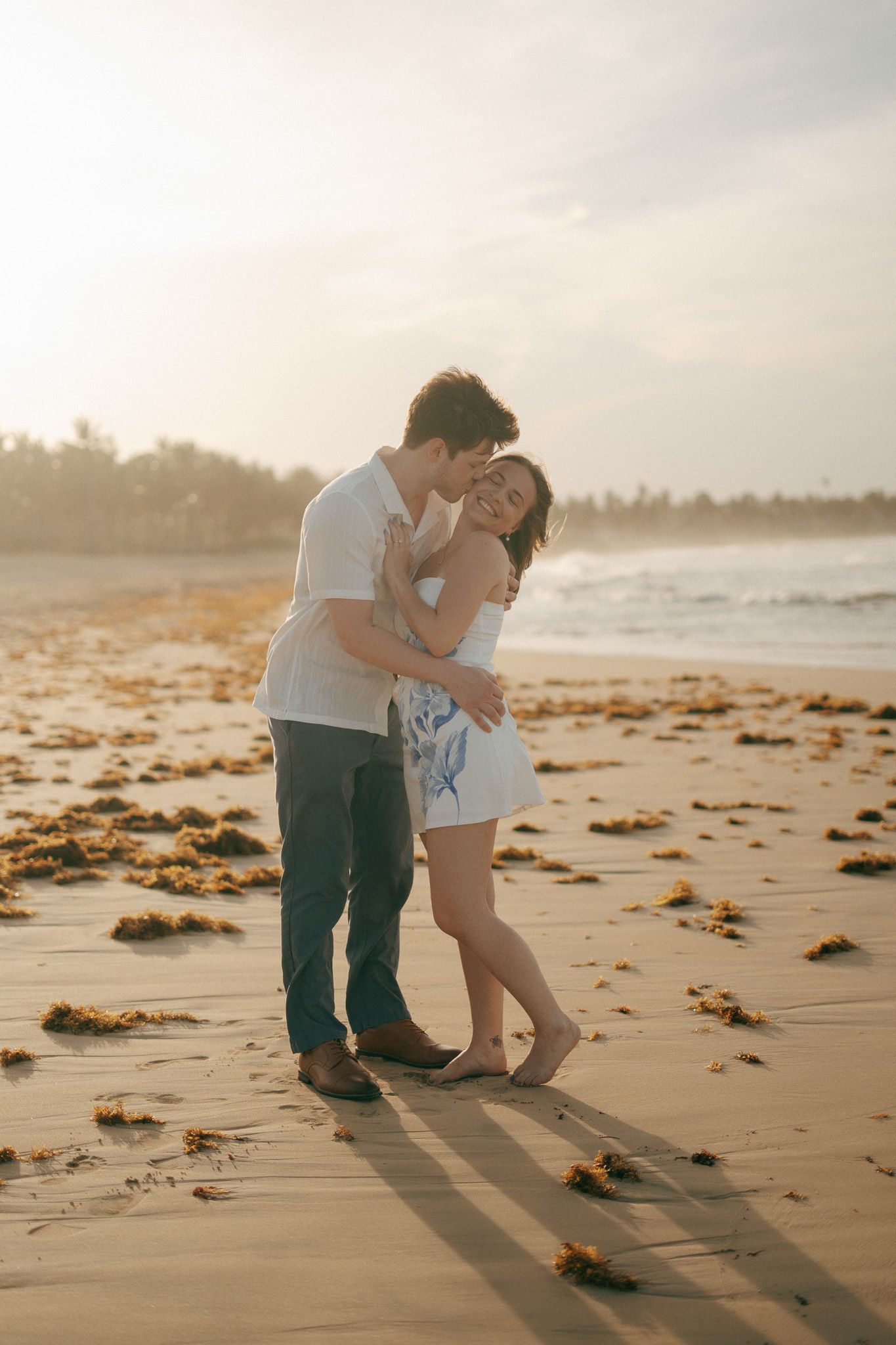 Couple embracing tenderly on a sunlit beach at sunset, man kissing woman's cheek as waves crash in the background.