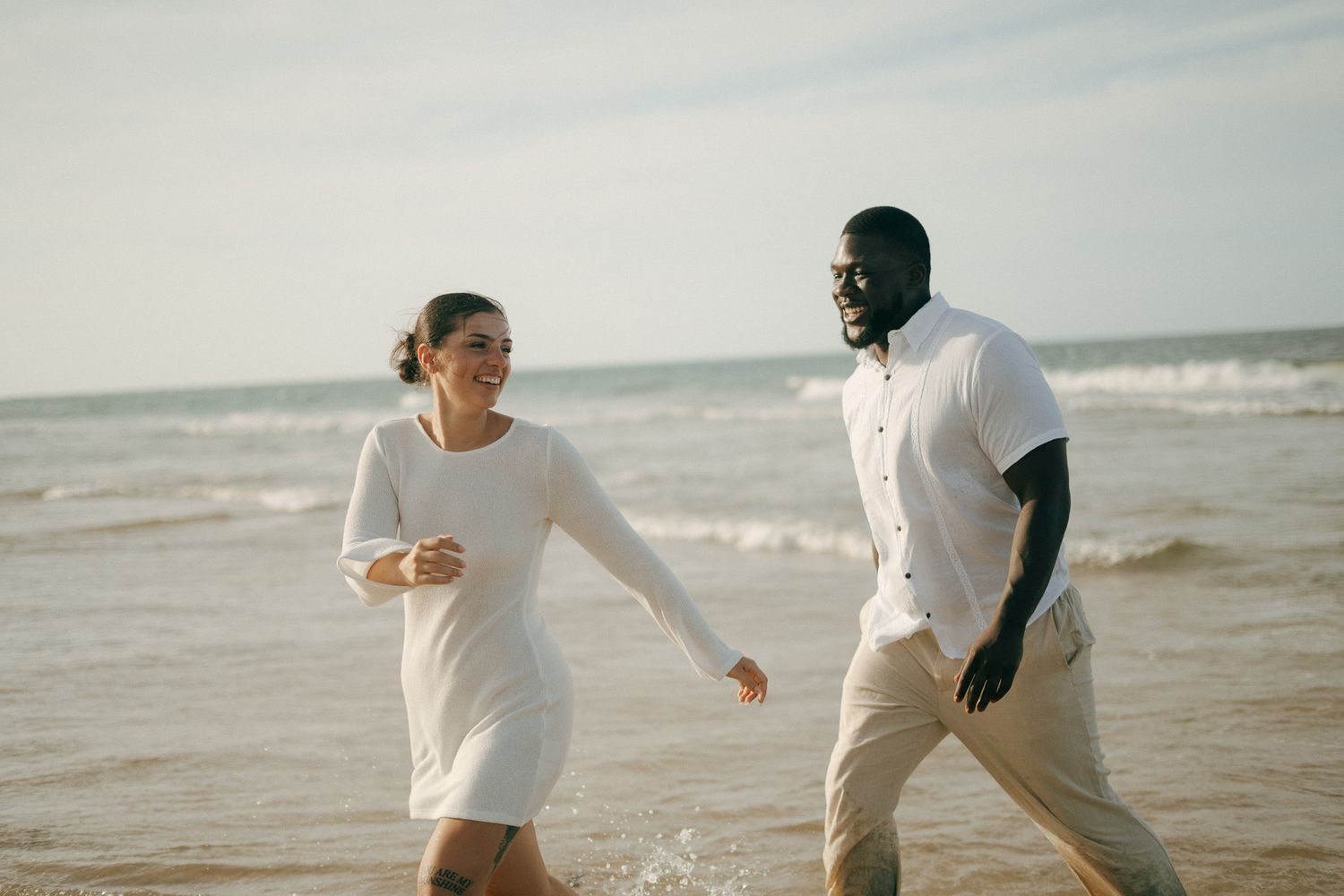 Happy interracial couple walking hand in hand on a sunny beach, smiling and enjoying a romantic seaside stroll.