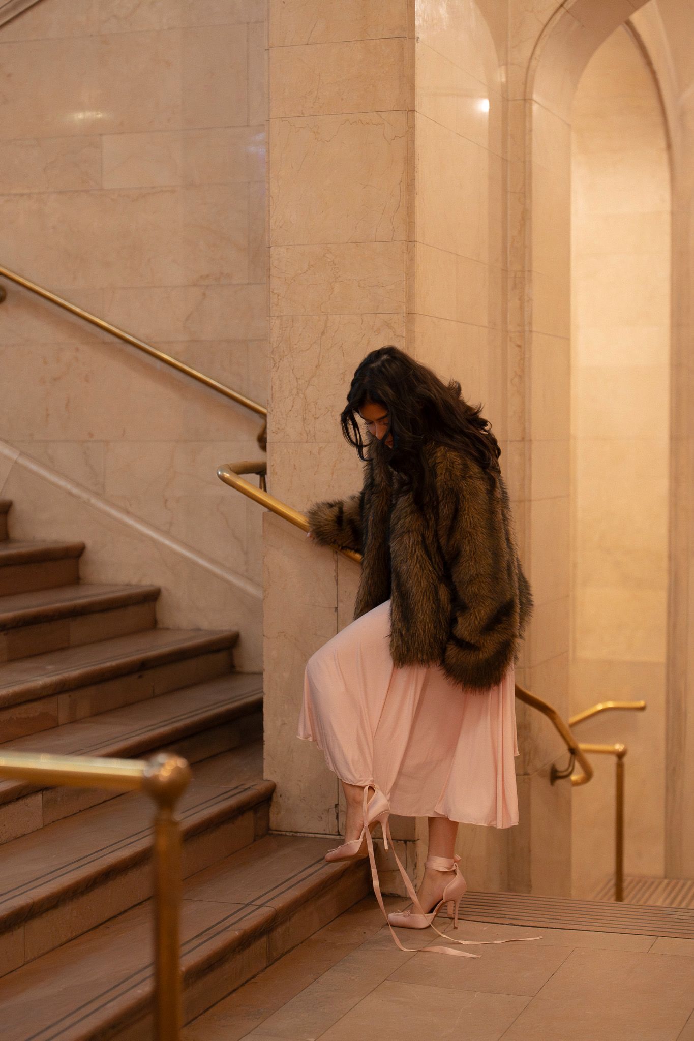 A figure in a pink dress and fur coat sits elegantly on marble stairs in a grand architectural setting.