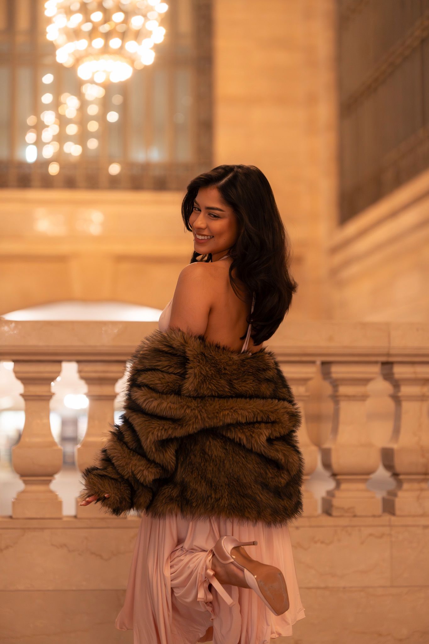 A woman in a brown fur wrap and pink dress poses elegantly against a grand marble balustrade with glowing lights.