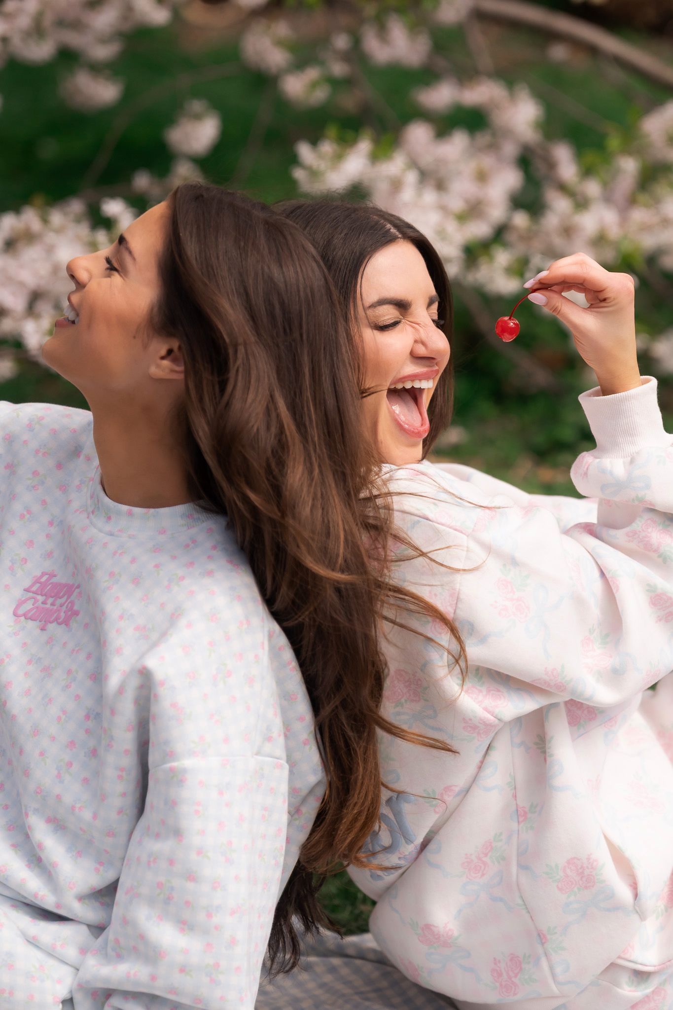 Two smiling women in matching pastel sweatshirts sit back-to-back outdoors near blooming flowers, one holding a cherry.