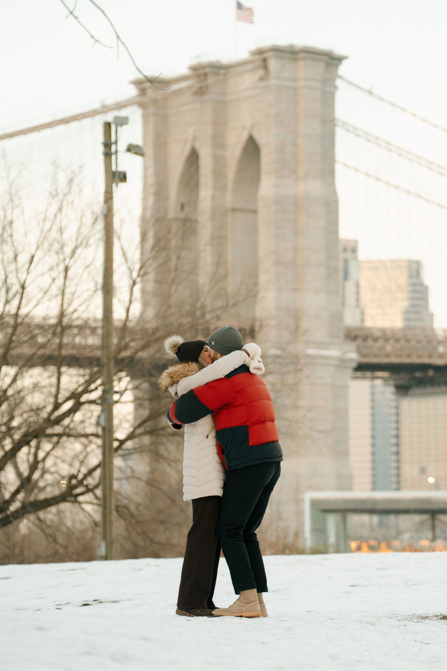 A couple embraces in the snow with the Brooklyn Bridge and city skyline visible in the background.