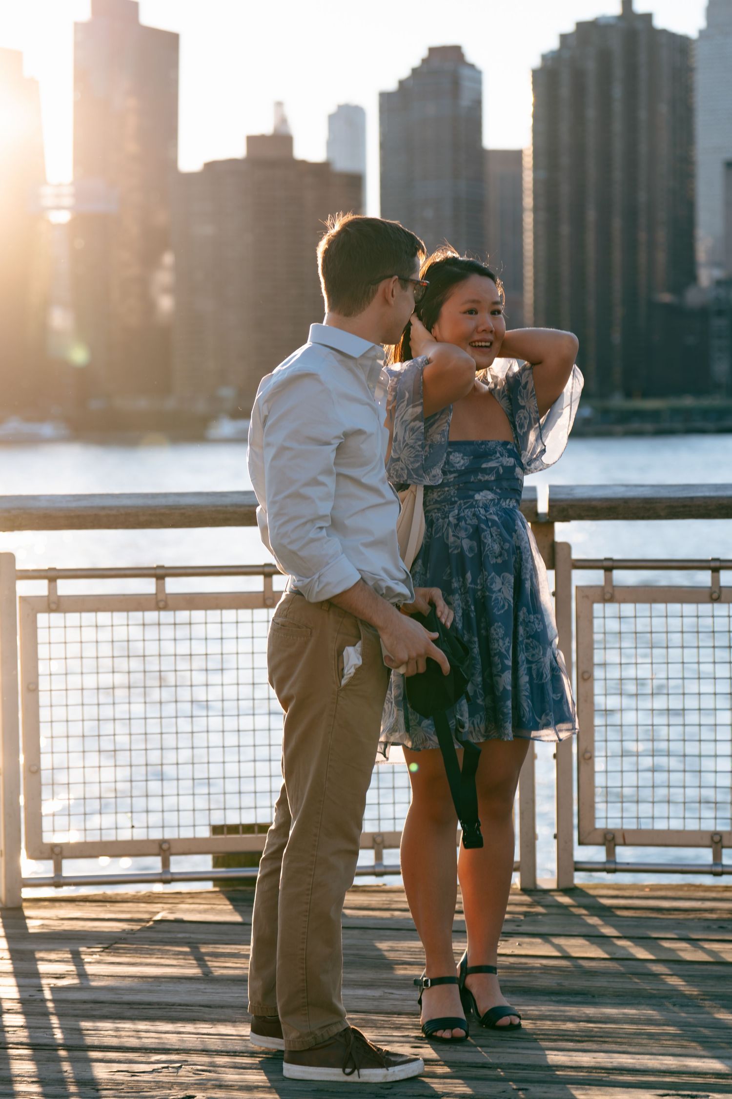A couple shares a romantic moment at sunset on a waterfront pier with city skyline in the background.