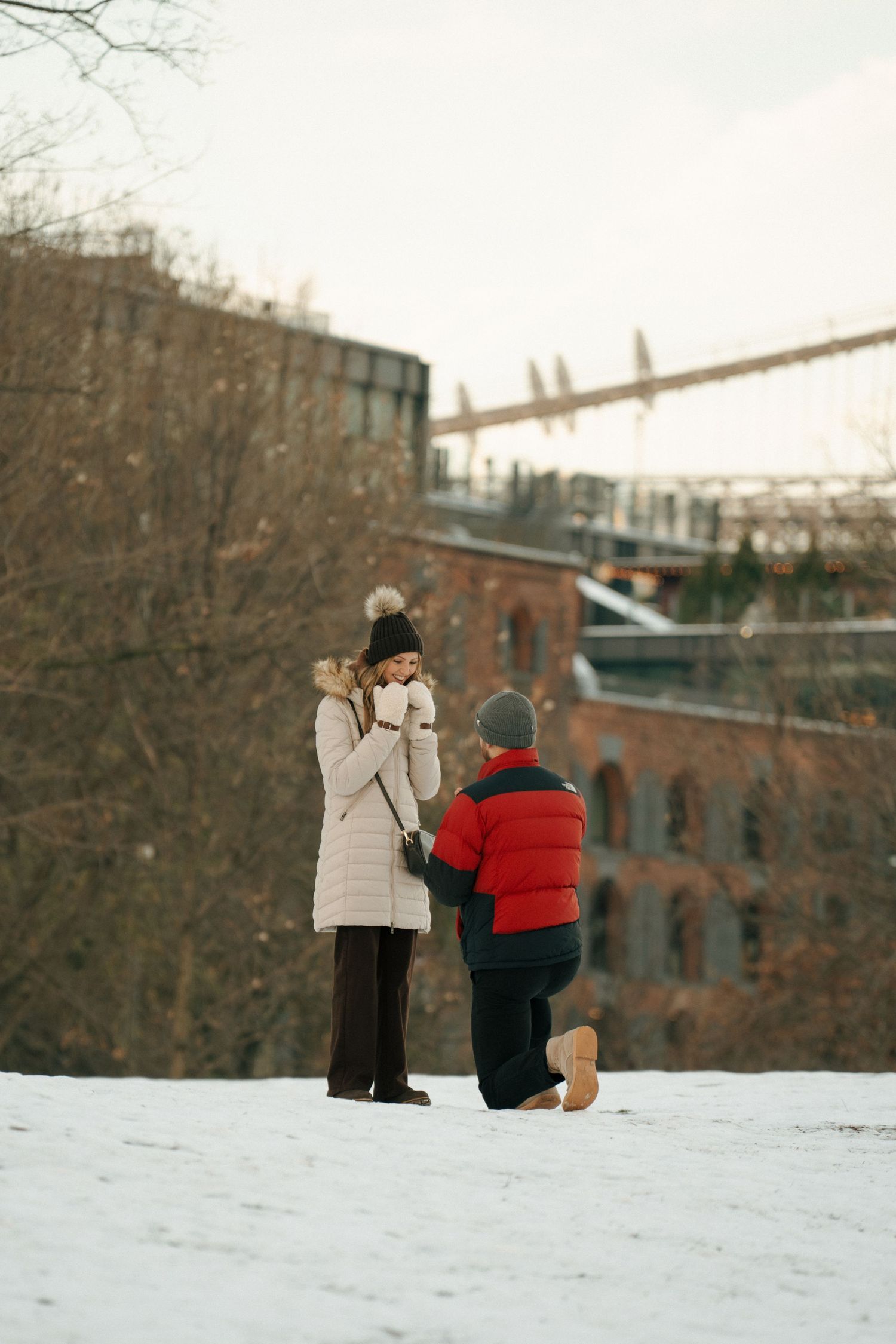Two people sit together in the snow on a winter day with brick buildings visible in the background.