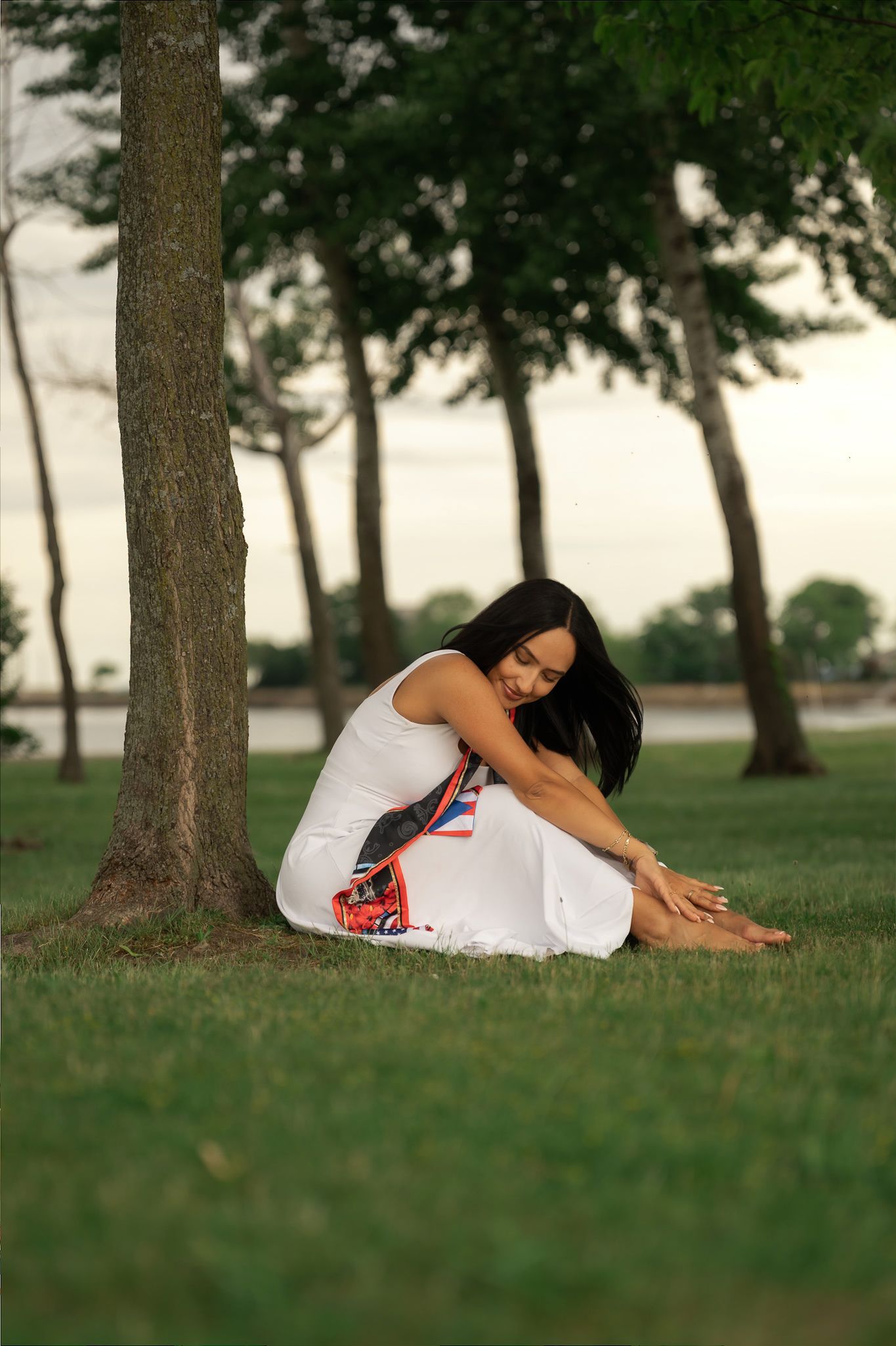 Woman in white dress sitting on grass leaning against tree in a serene park setting at dusk.