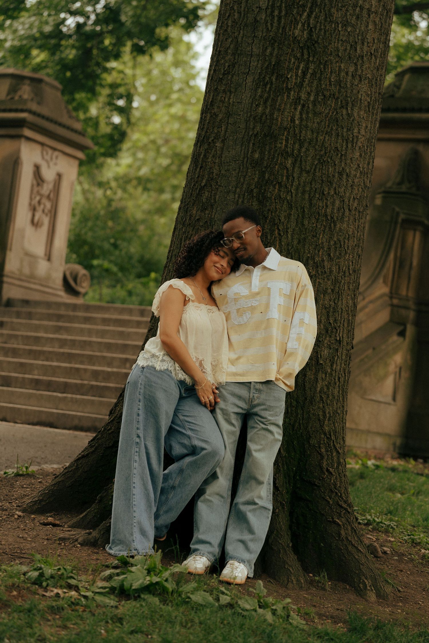 A couple leans against a large tree in a park, sharing a tender moment in casual denim outfits.