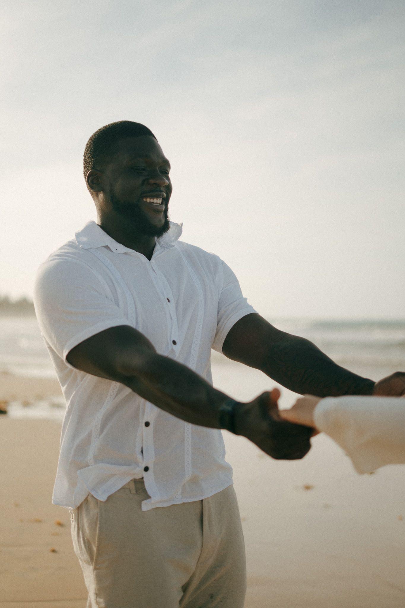 Smiling man in white shirt enjoying a beach day, reaching out with open arms on sandy shore.