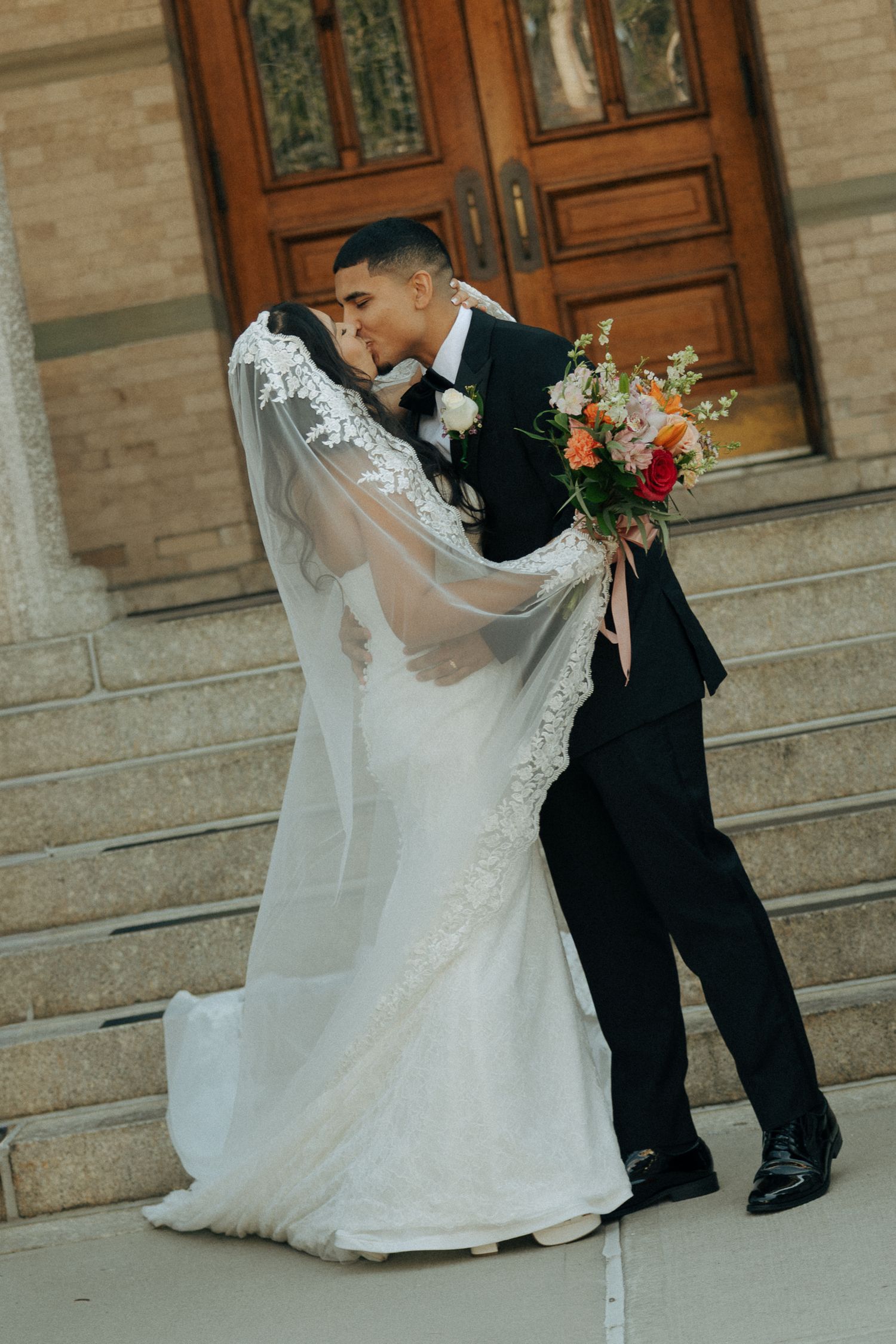 Bride and groom share a romantic kiss on church steps, bride in lace wedding gown with veil holding colorful bouquet.