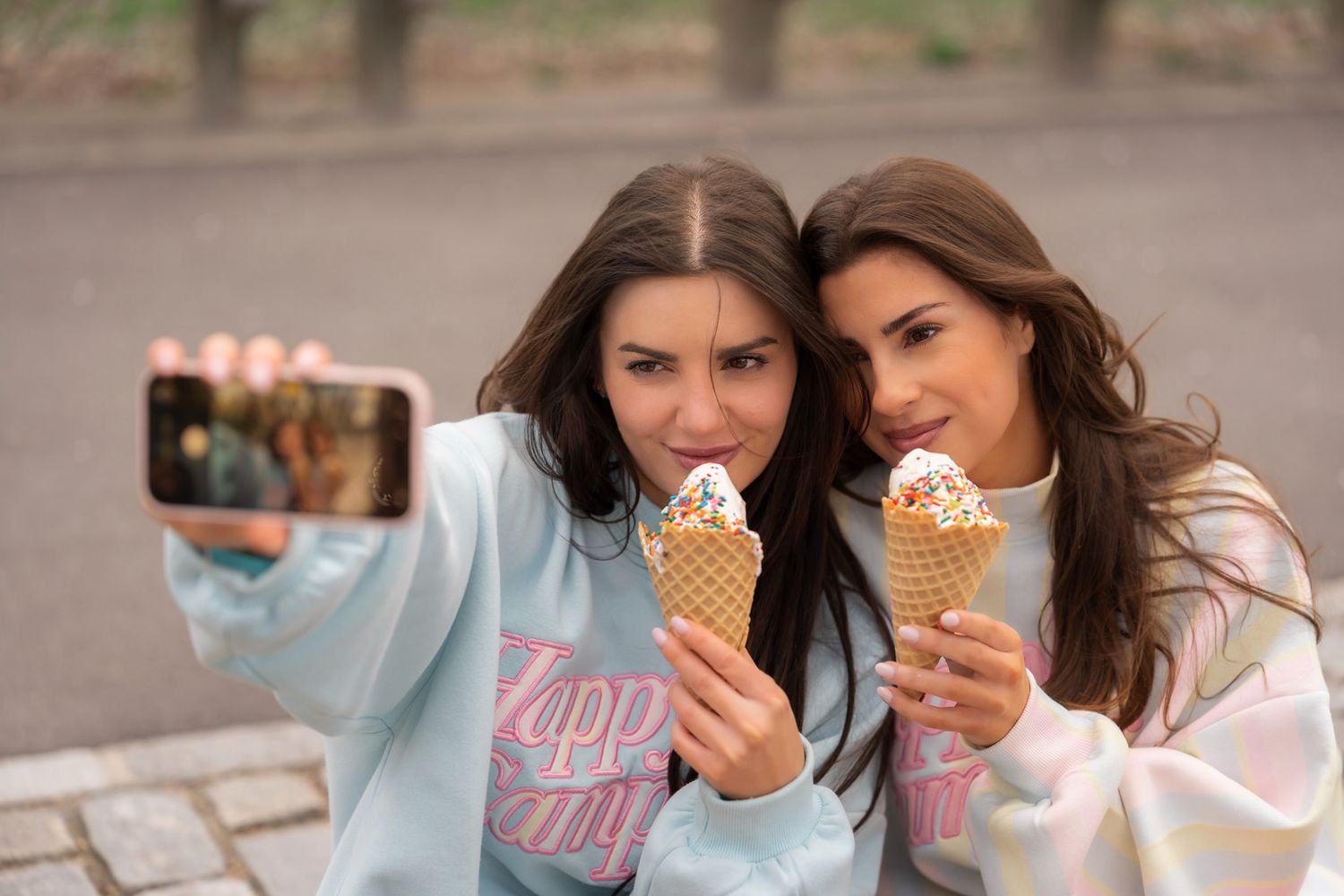 Two women taking a selfie while holding ice cream cones, wearing matching Happy Camper graphic sweatshirts.