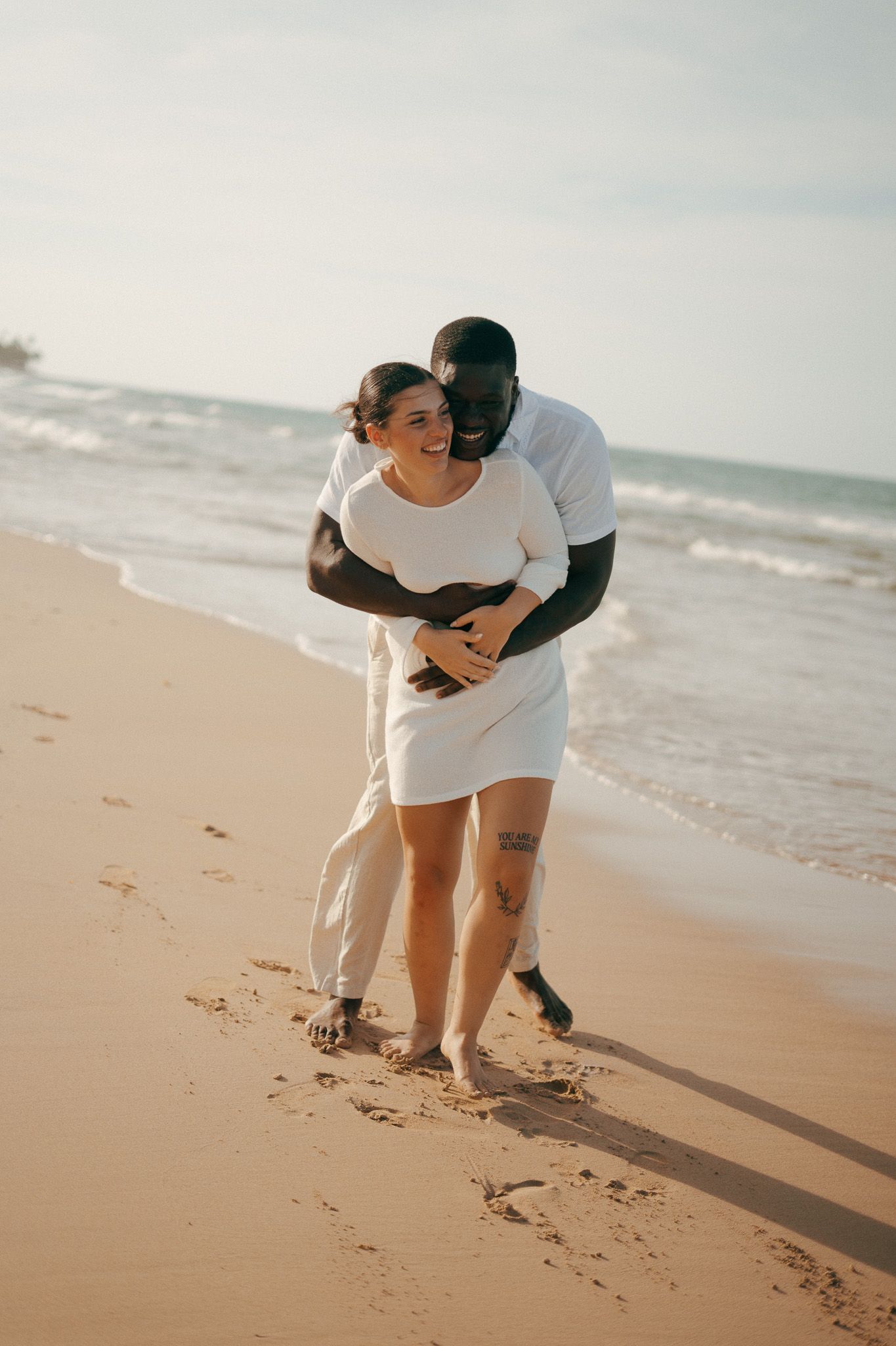 Happy interracial couple embracing and walking barefoot on a sandy beach near ocean waves in white outfits.
