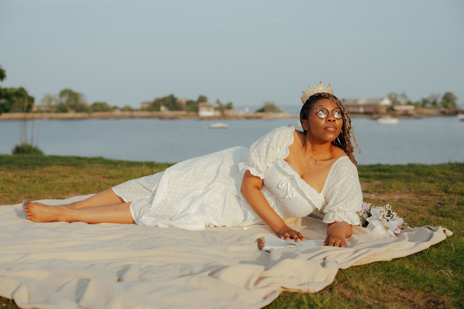 Person in white dress lying on grass against a coastal background at sunset.