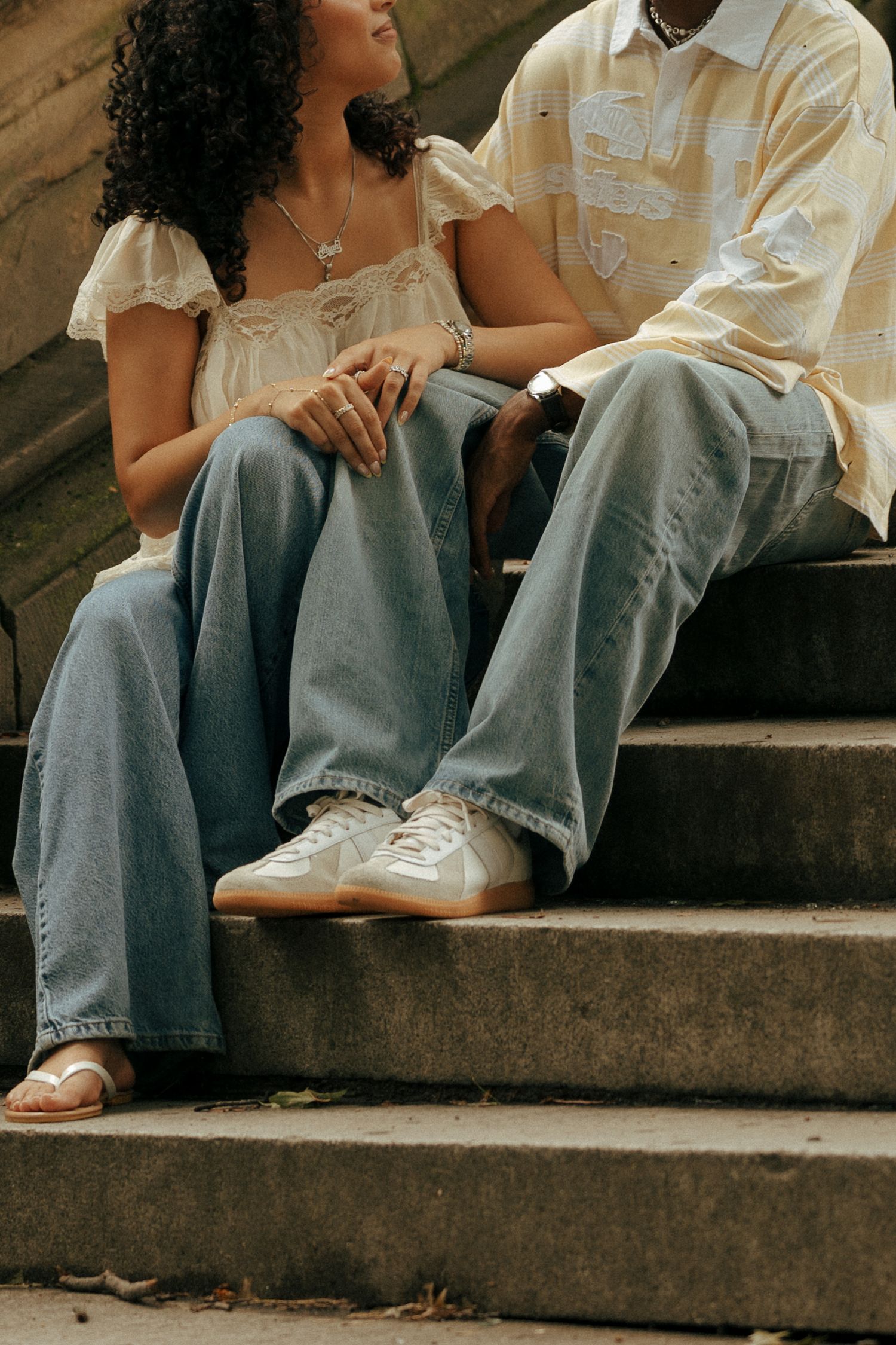 Couple sitting on stone steps wearing wide-leg jeans, white sneakers, and casual tops in an outdoor setting.