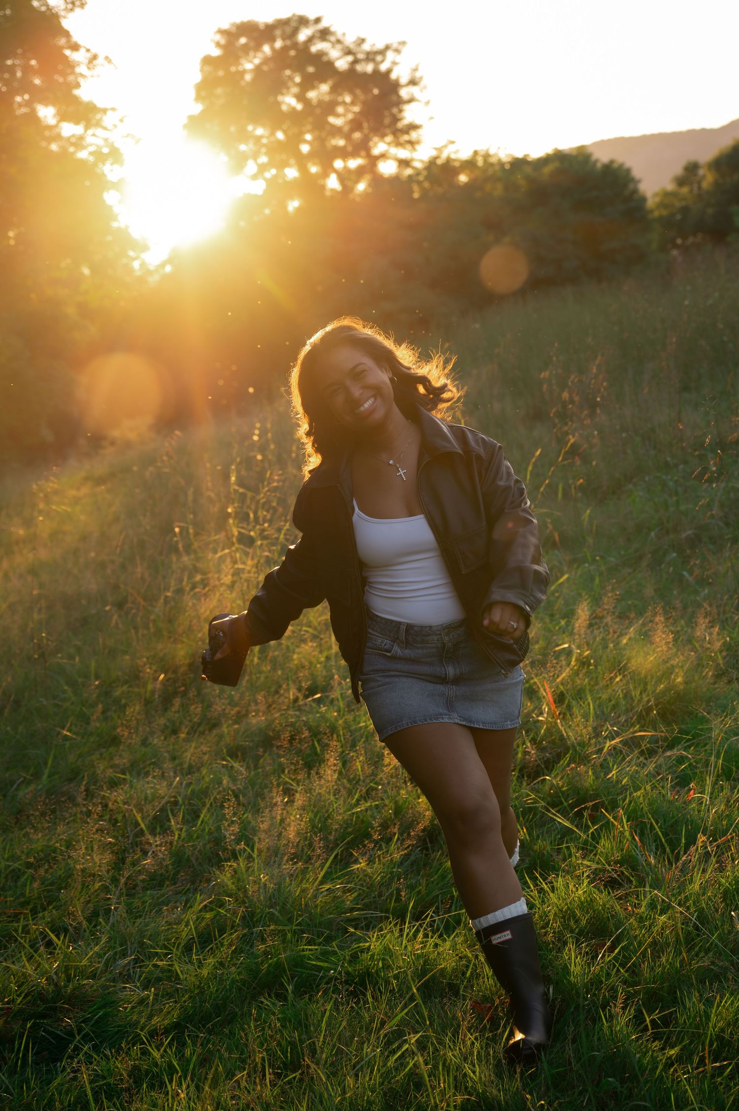 Young woman dancing in a sunlit field at golden hour, wearing a denim skirt, white top, and Hunter boots.