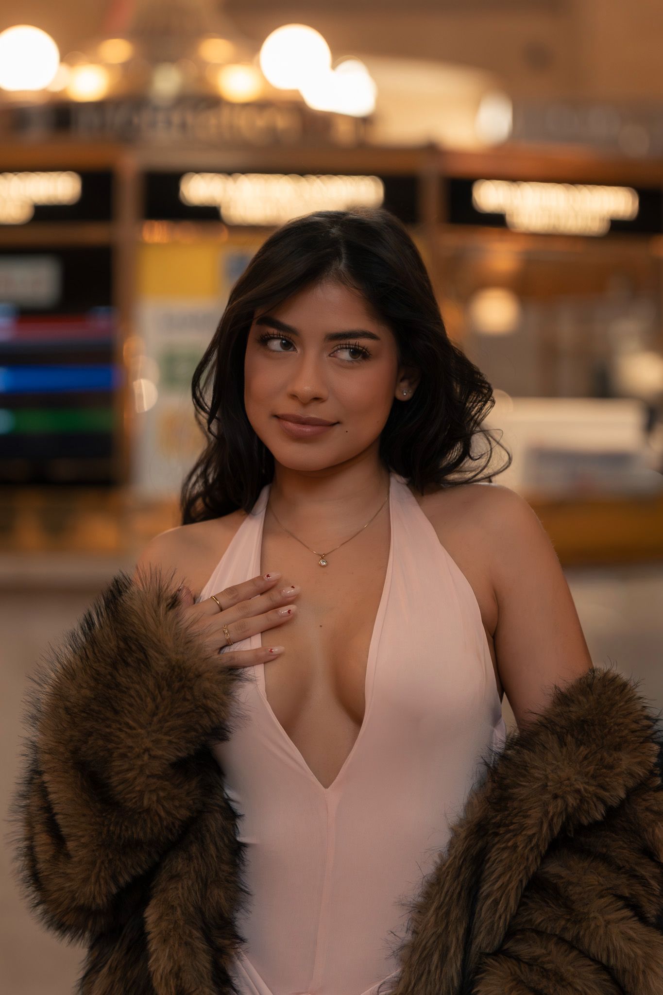 A woman in a blush pink bodysuit poses with a brown fur coat in an elegant indoor setting.