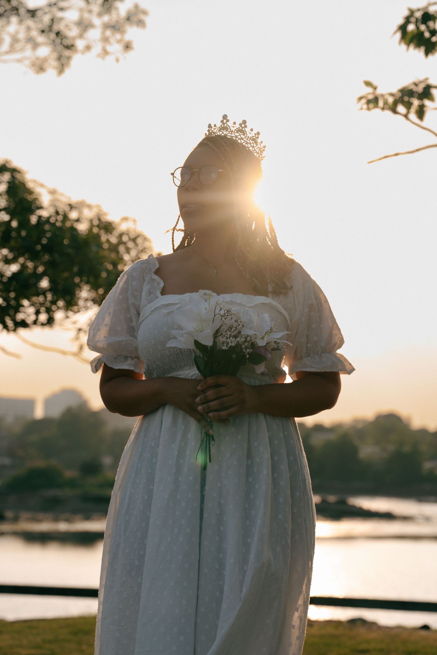 Person in white dress holding flowers against sunset over water.