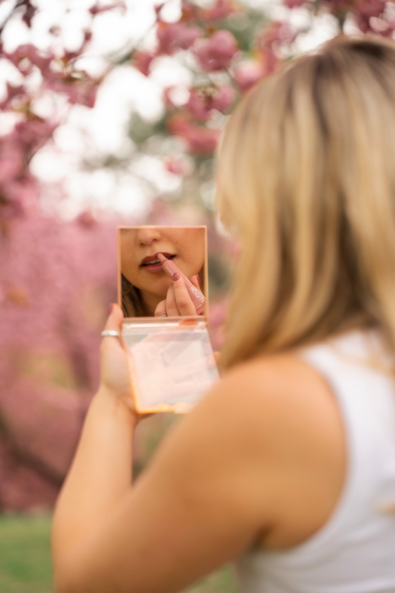 Blonde woman applying lipstick using a compact mirror outdoors near pink cherry blossom trees.