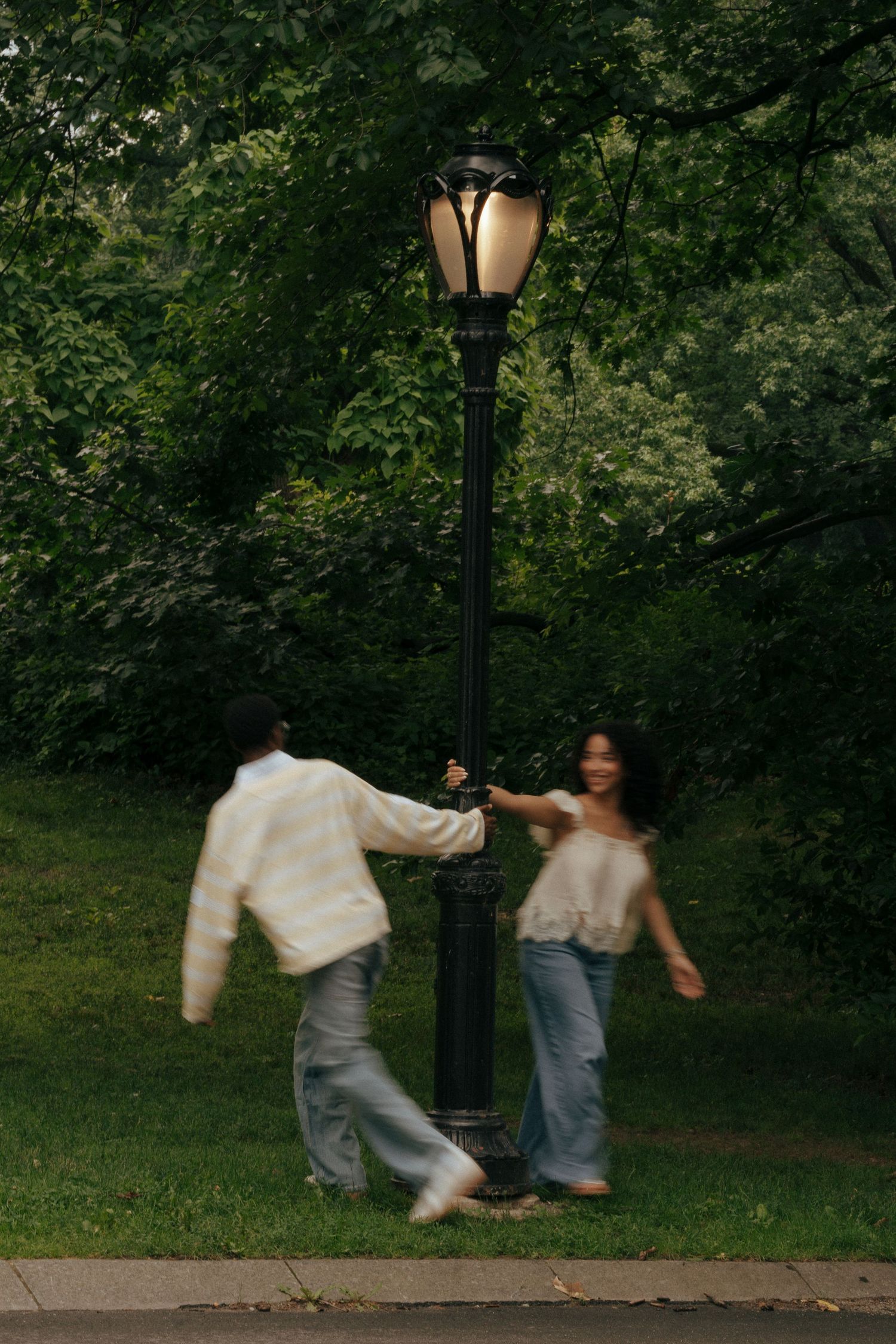 Couple playfully spinning around a glowing park lamp post surrounded by lush green trees.
