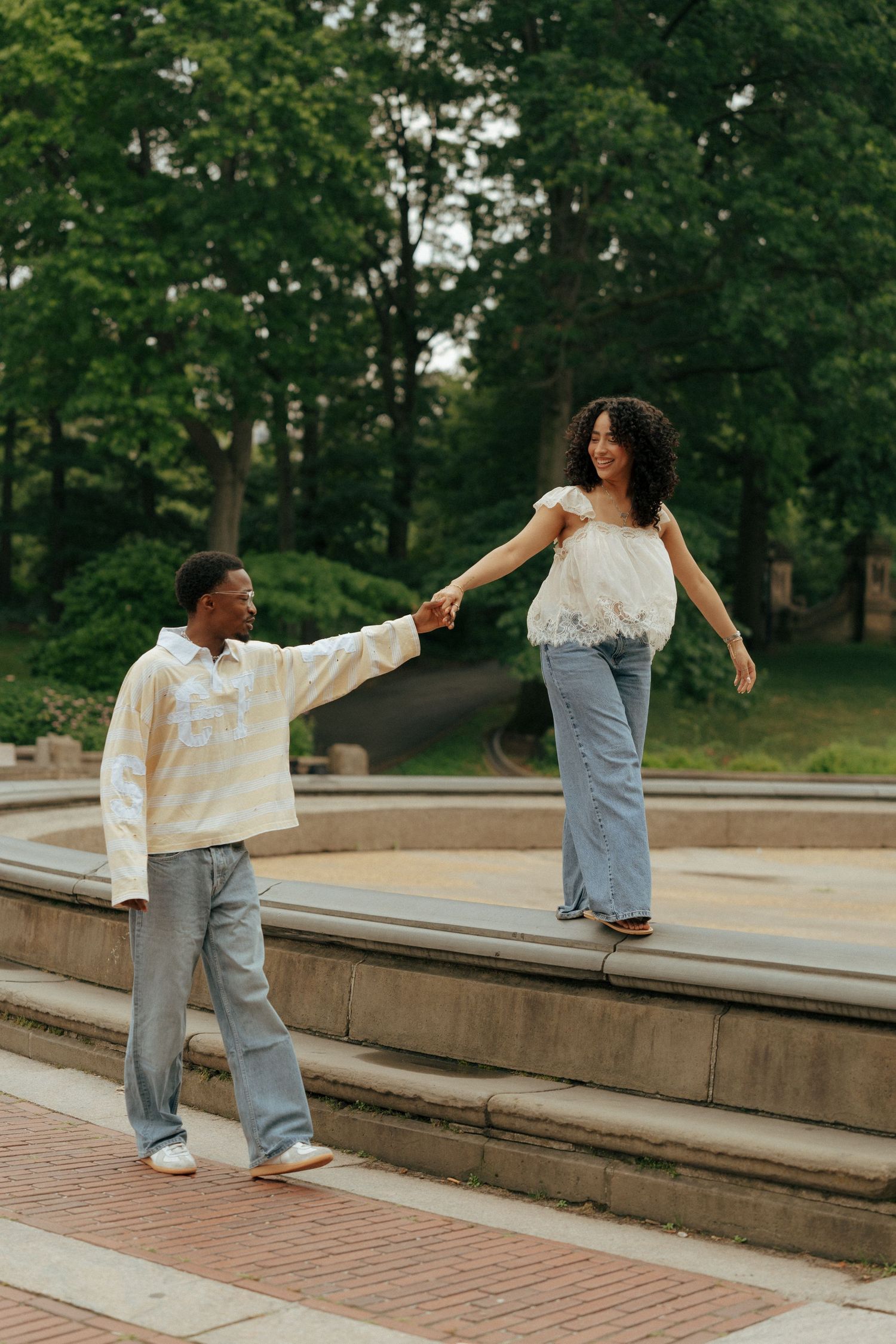 A young couple holds hands in a lush green park, the woman balancing on a ledge while smiling joyfully.