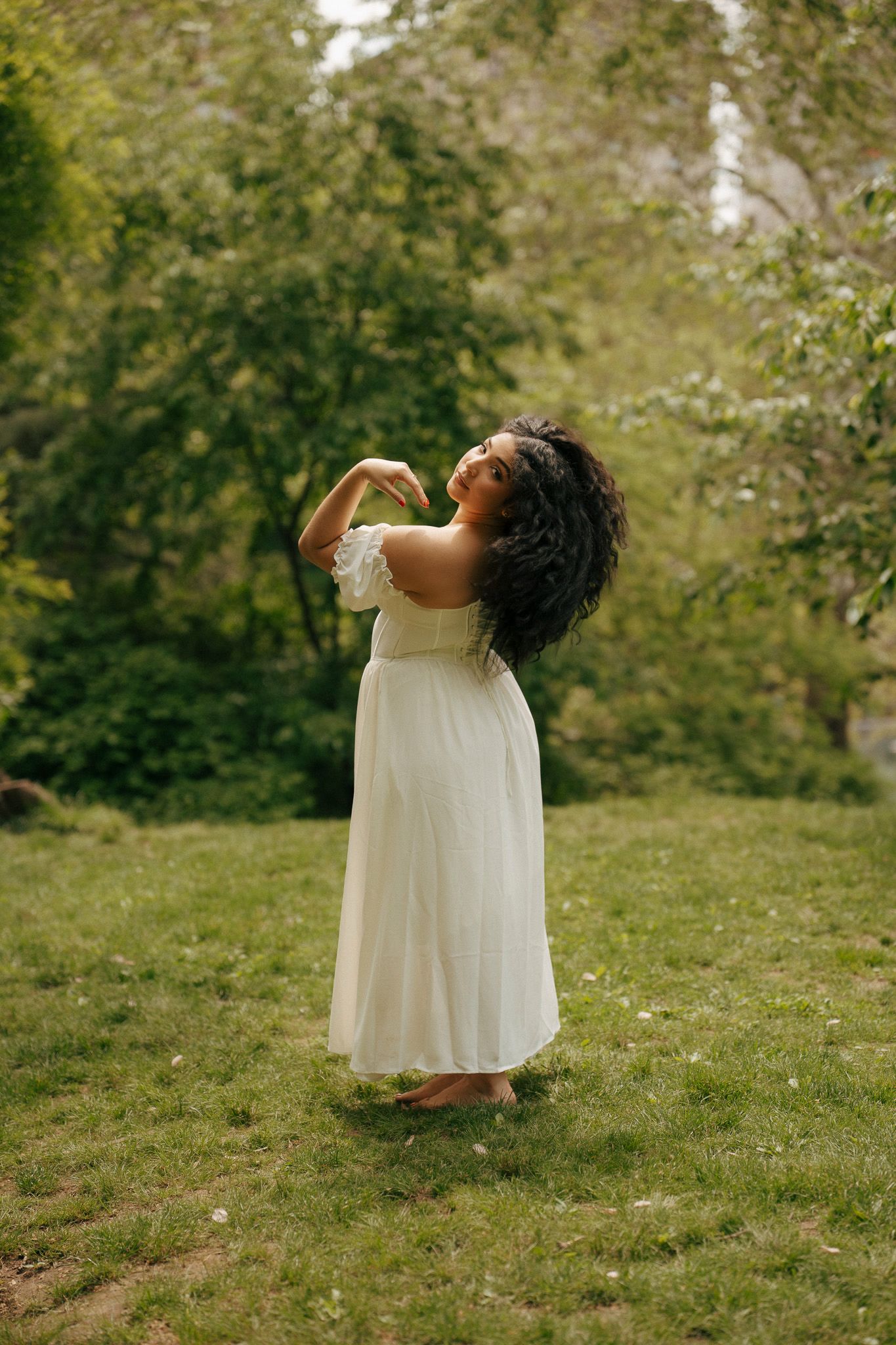 Woman in white off-shoulder dress dancing barefoot on green grass surrounded by lush garden trees.