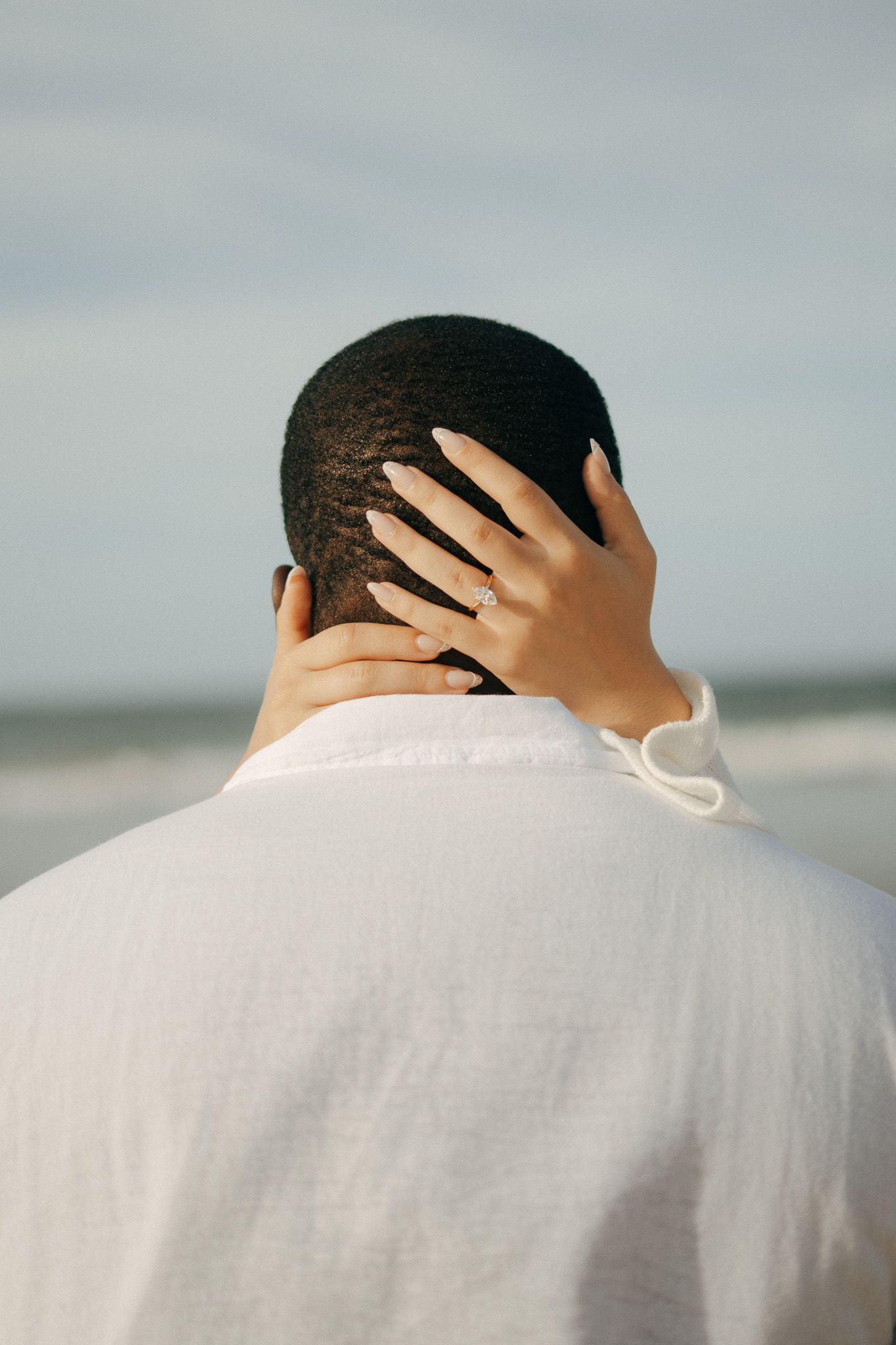 A woman's hand with an engagement ring rests on a man's head at the beach.