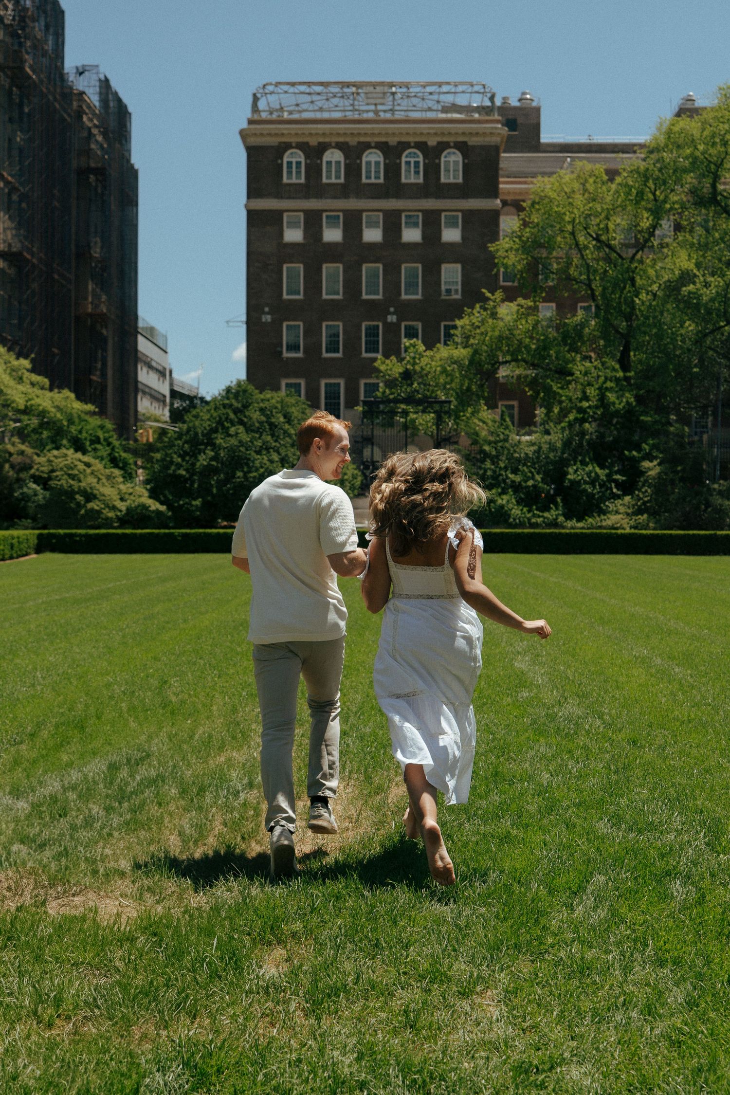 Couple in white clothing running playfully on a green lawn near brick buildings.