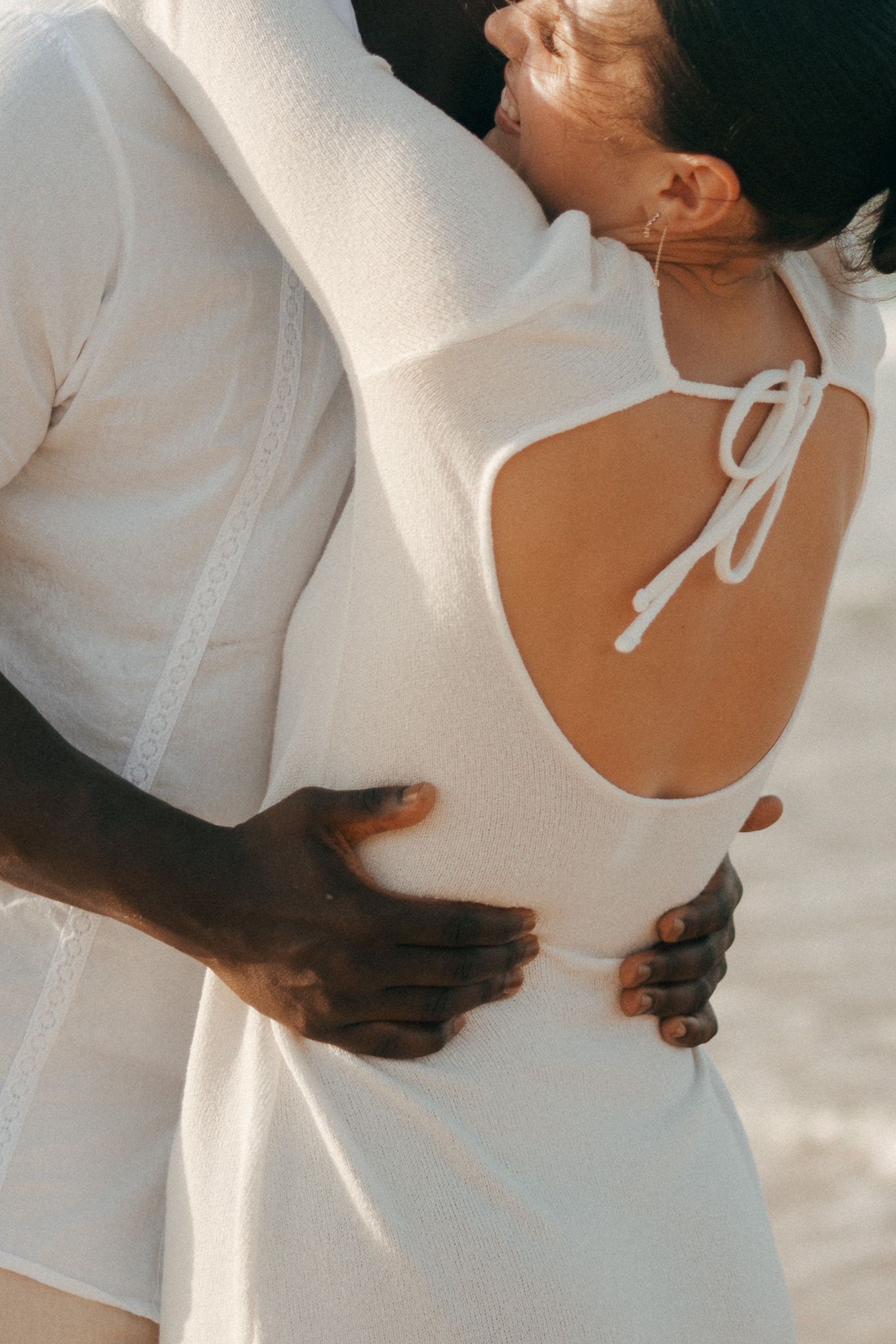 Close-up of an interracial couple embracing, woman in white open-back dress with bow tie detail at beach.