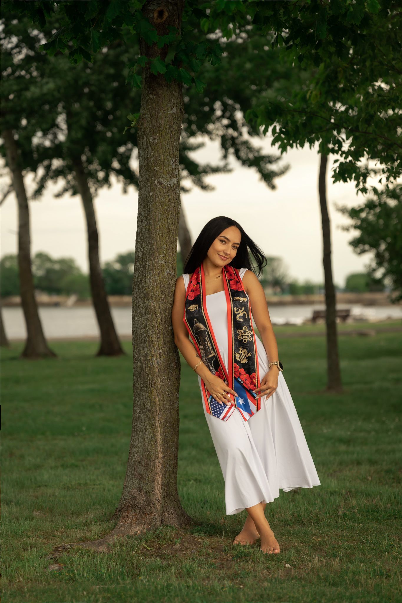 Young woman in white dress and colorful sash leans against tree in park, barefoot on green grass.