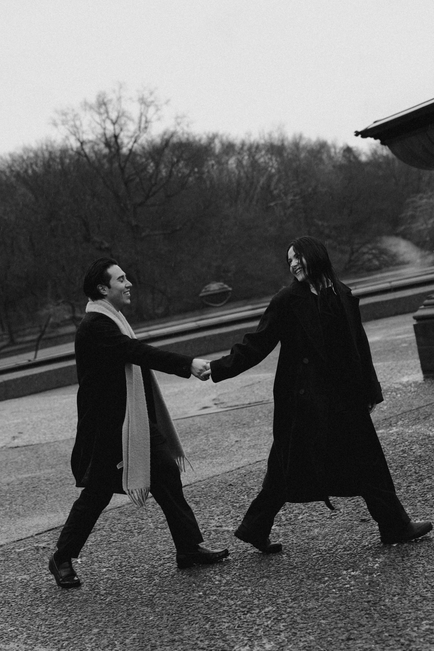 Two people in black coats playfully engage in a choreographed dance-like sequence outdoors on a grassy hill.