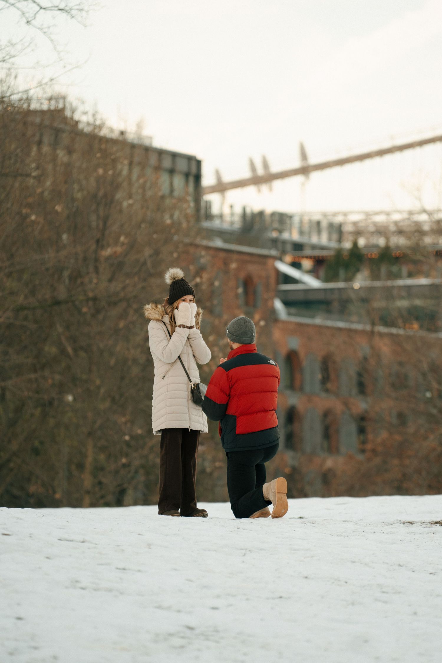 Two people in winter coats stand together in the snow with brick buildings and industrial structures in the background.