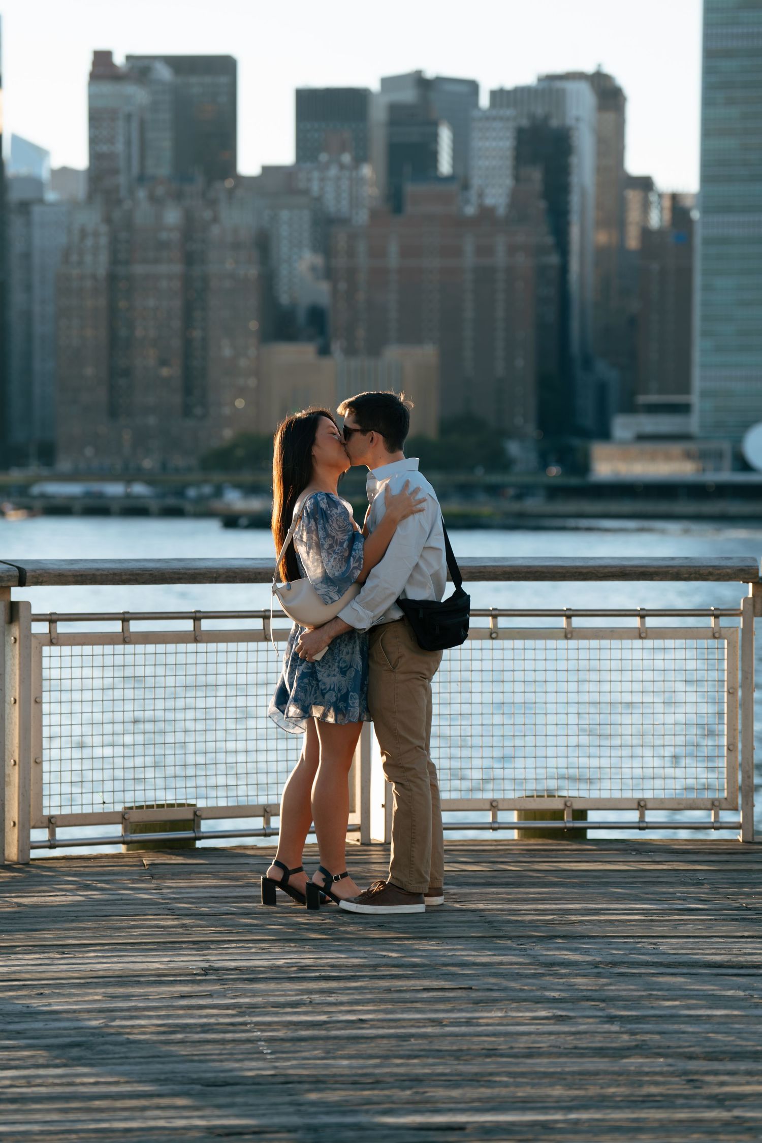 A romantic couple shares an intimate moment on a waterfront pier with the New York City skyline in the background.