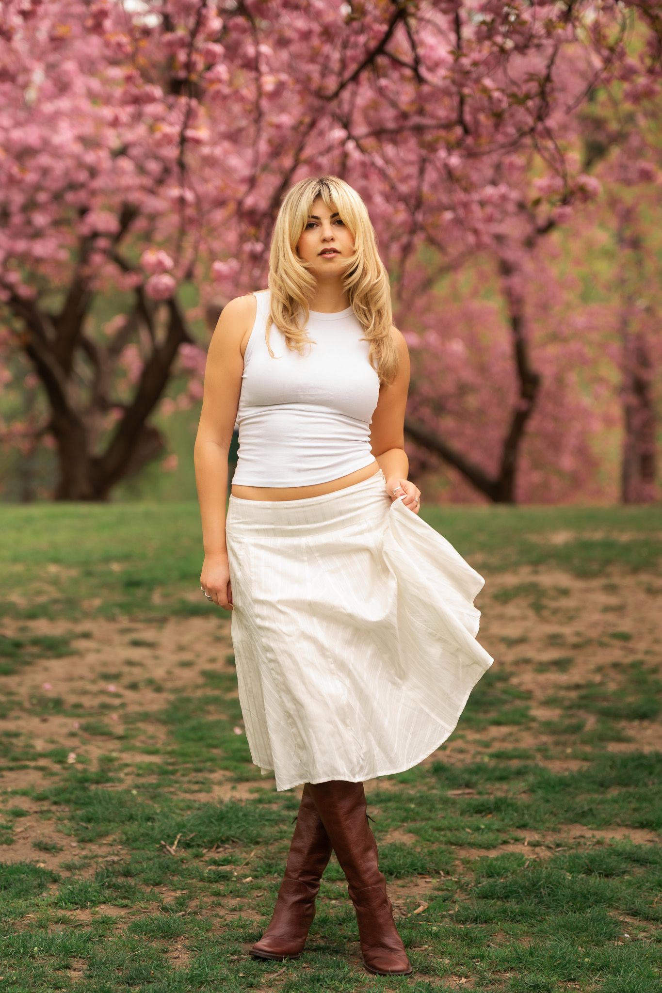 Blonde woman in white crop top, skirt, and brown boots standing in a park with pink cherry blossom trees.