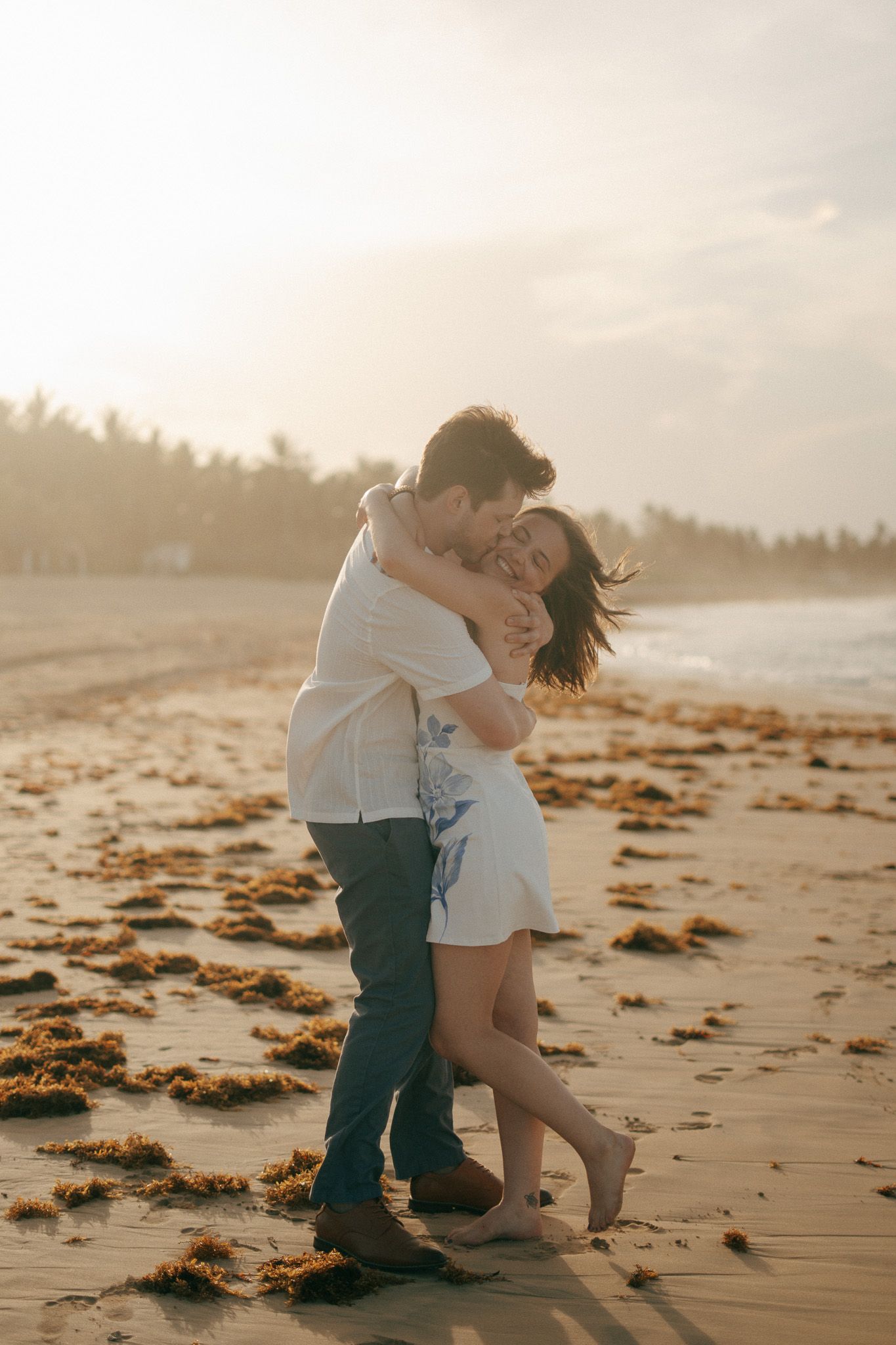Couple embracing romantically on a sandy beach at golden sunset, woman in white dress smiling joyfully, palm trees in background.