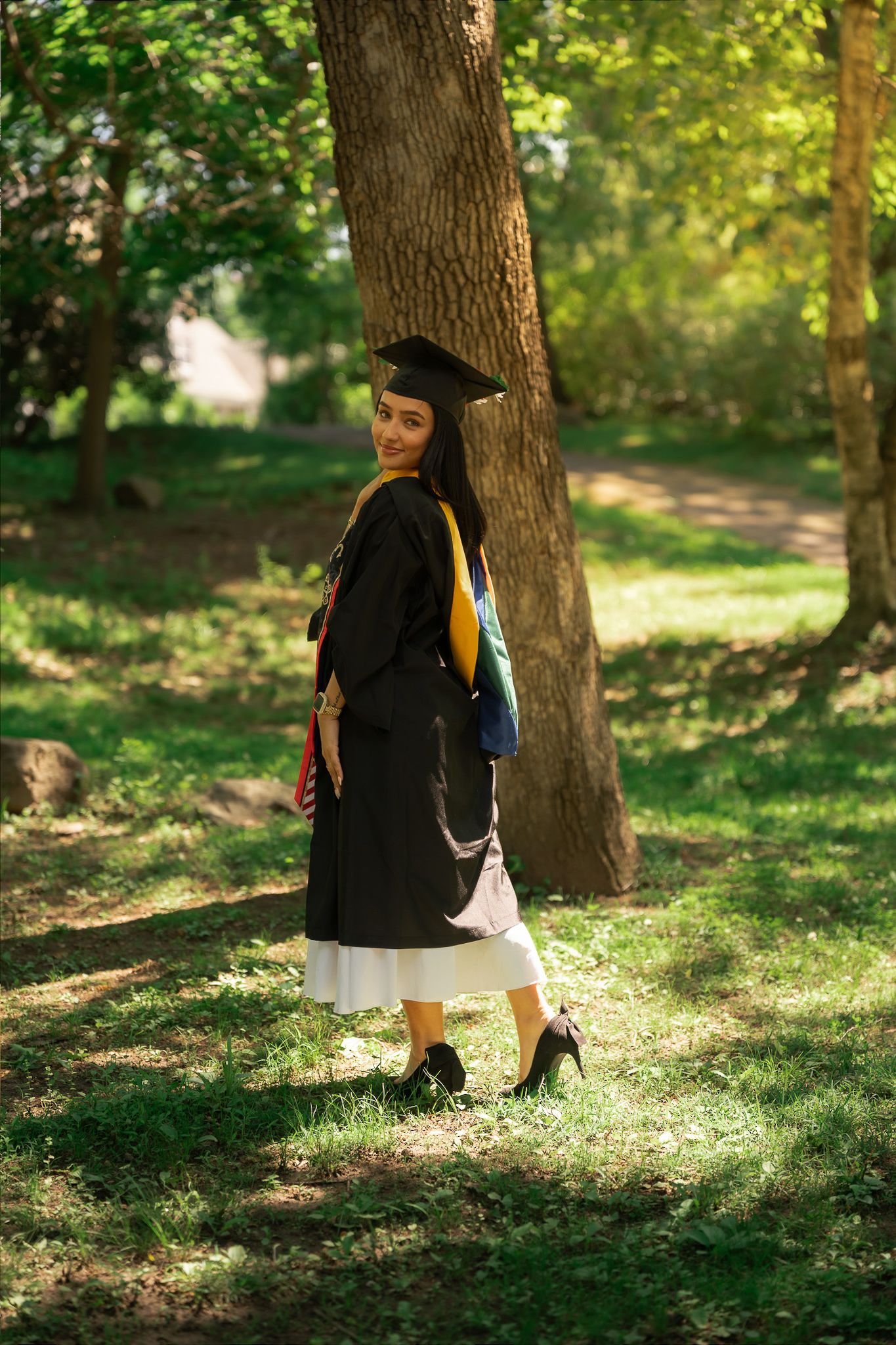 A young woman in black graduation gown and cap stands outdoors in a sunny green park, smiling over her shoulder.