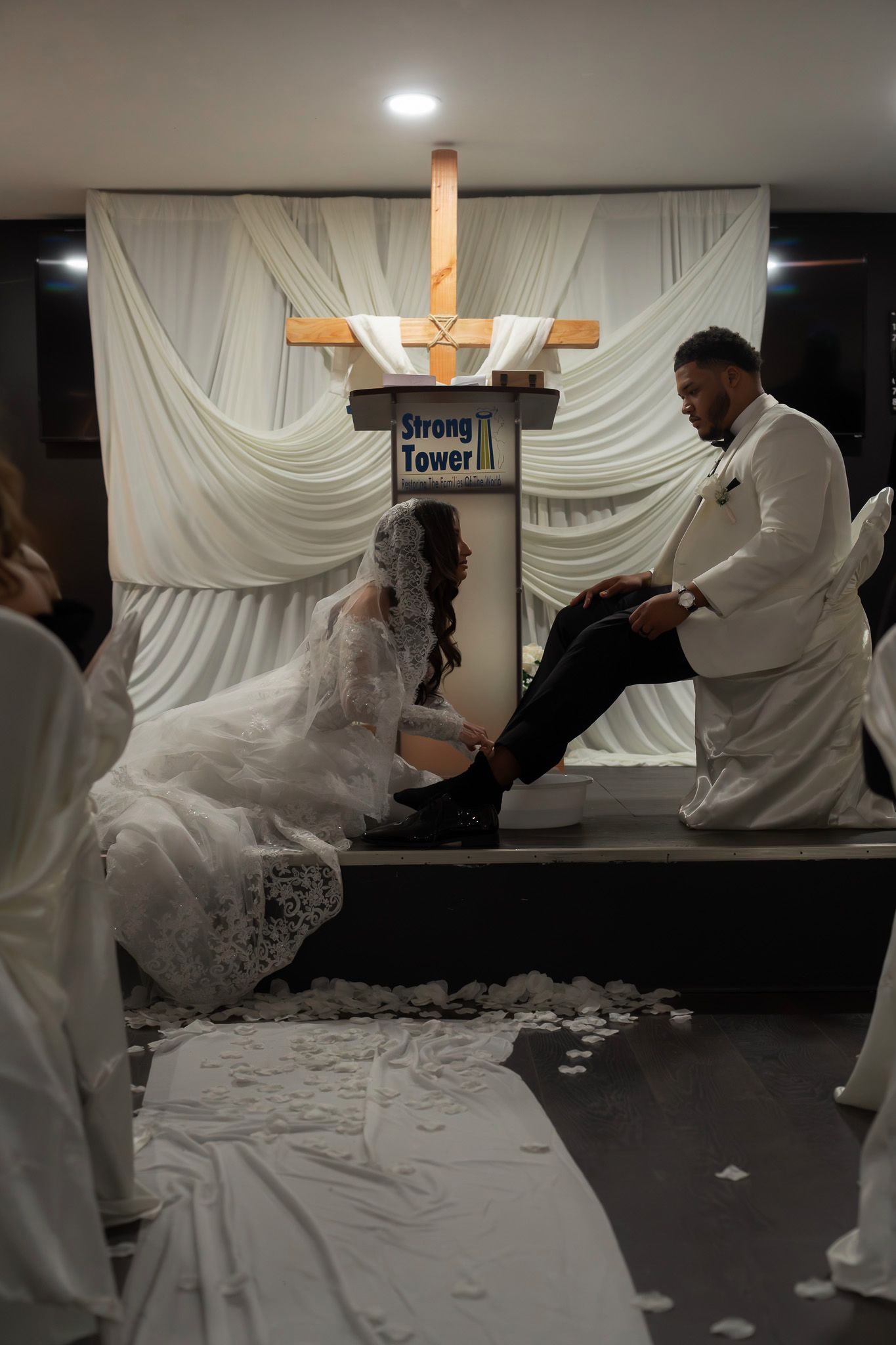 A dramatic scene of a wedding ceremony with people in white attire gathered around a wooden cross on stage.