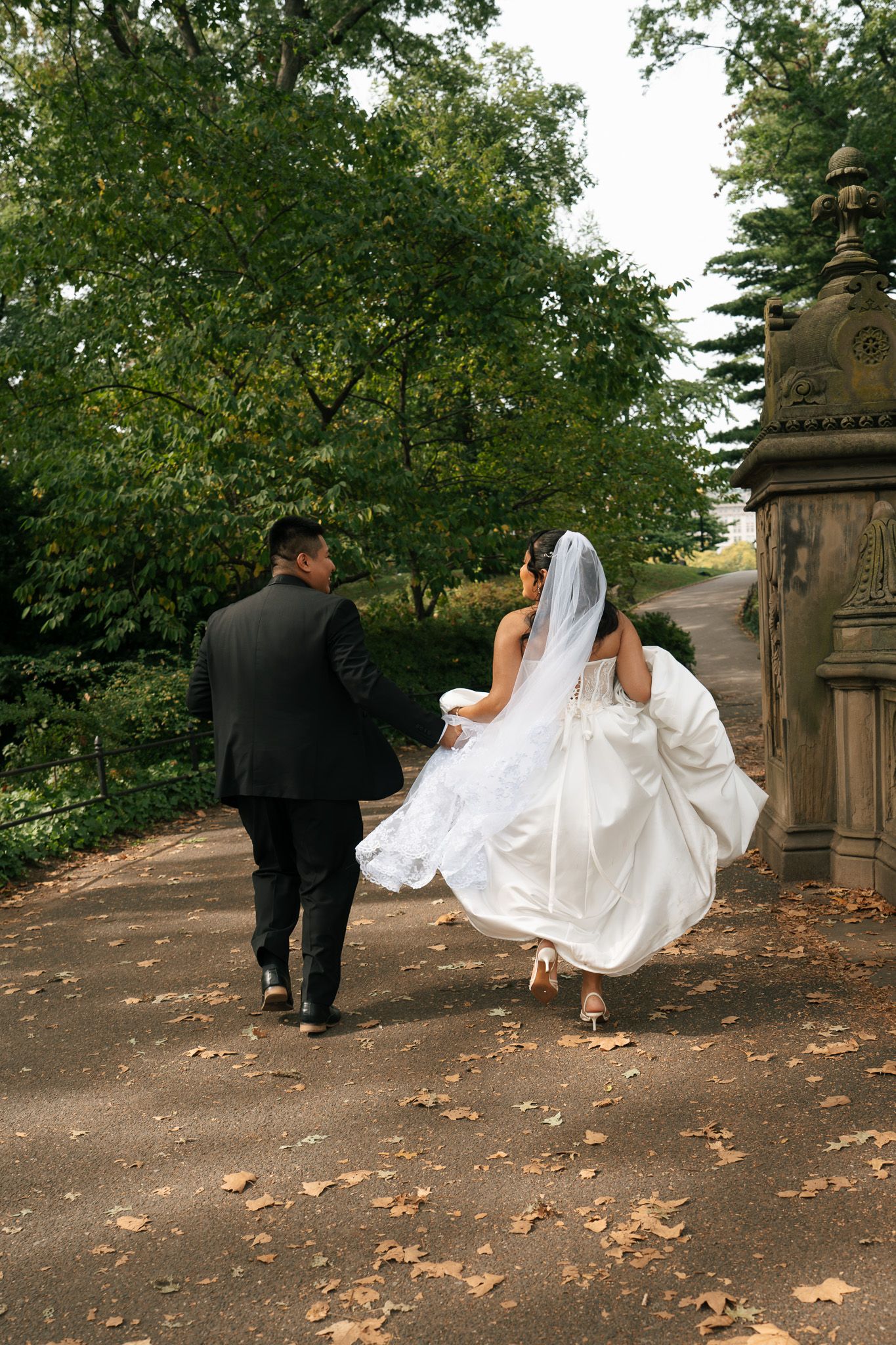 A couple in wedding attire walks along a tree-lined path with autumn leaves scattered on the ground.