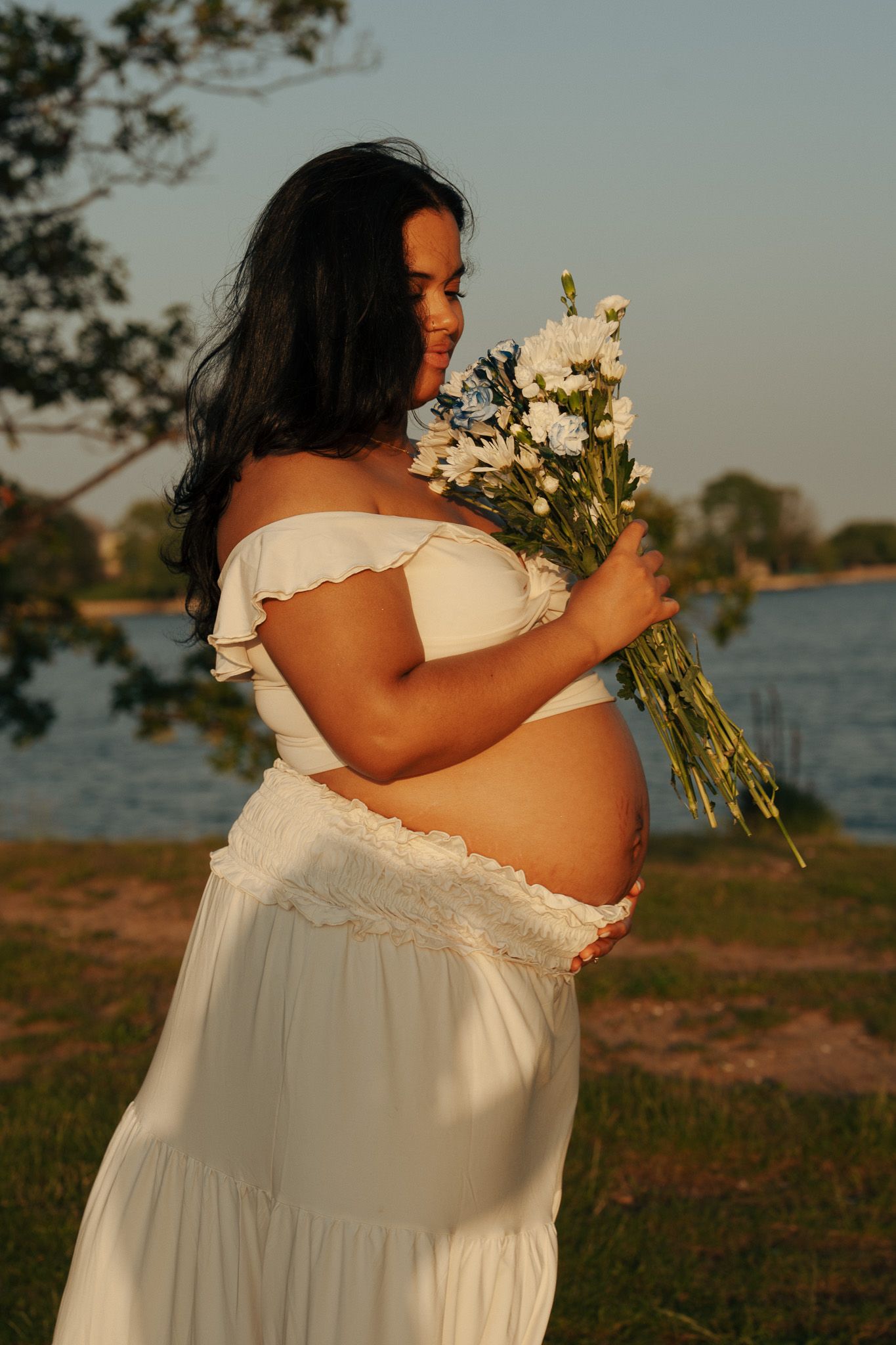 Expectant mother in flowing white dress holds flowers against sunset waterfront backdrop.