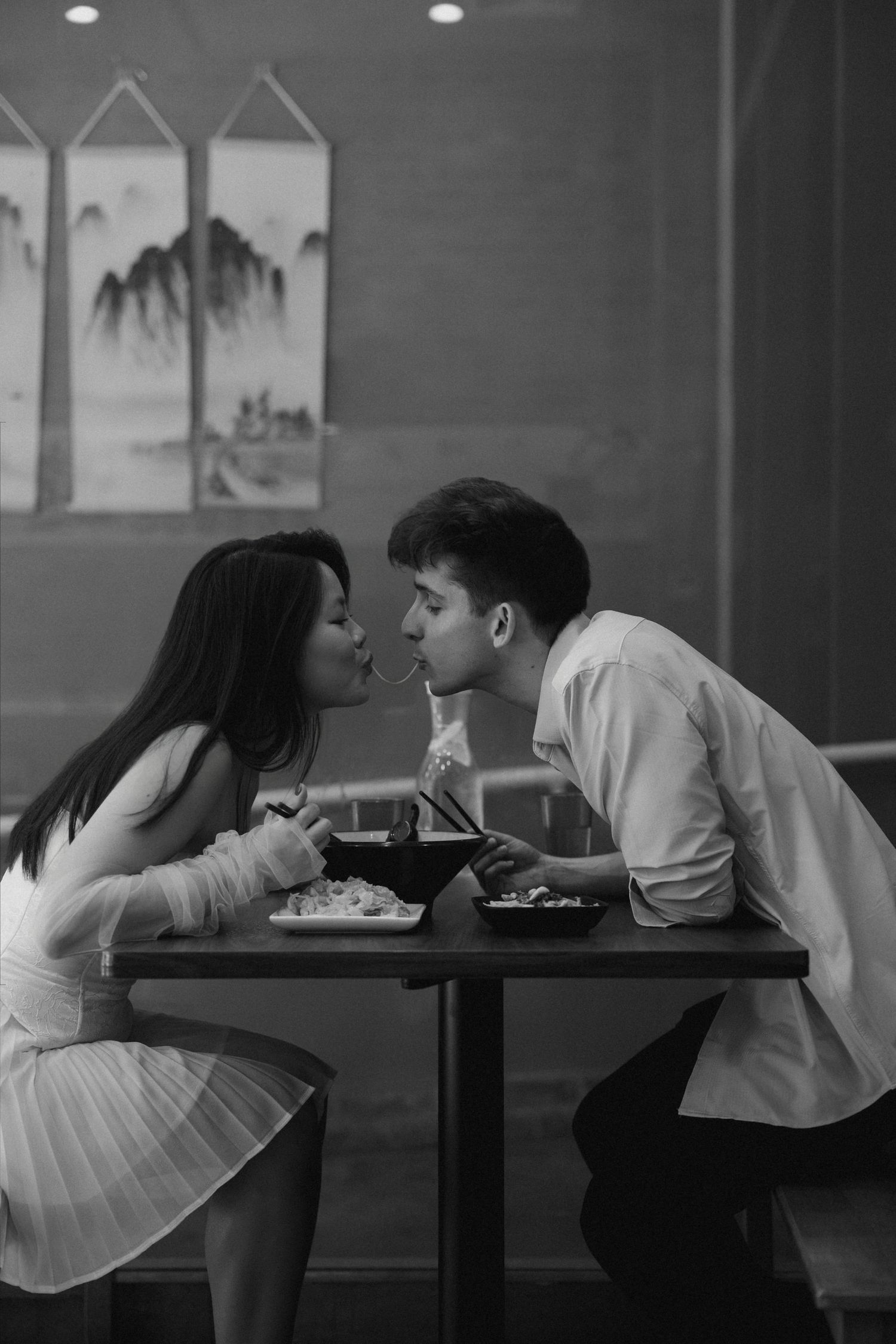 A couple shares a romantic noodle kiss at a restaurant table in this black and white photo.