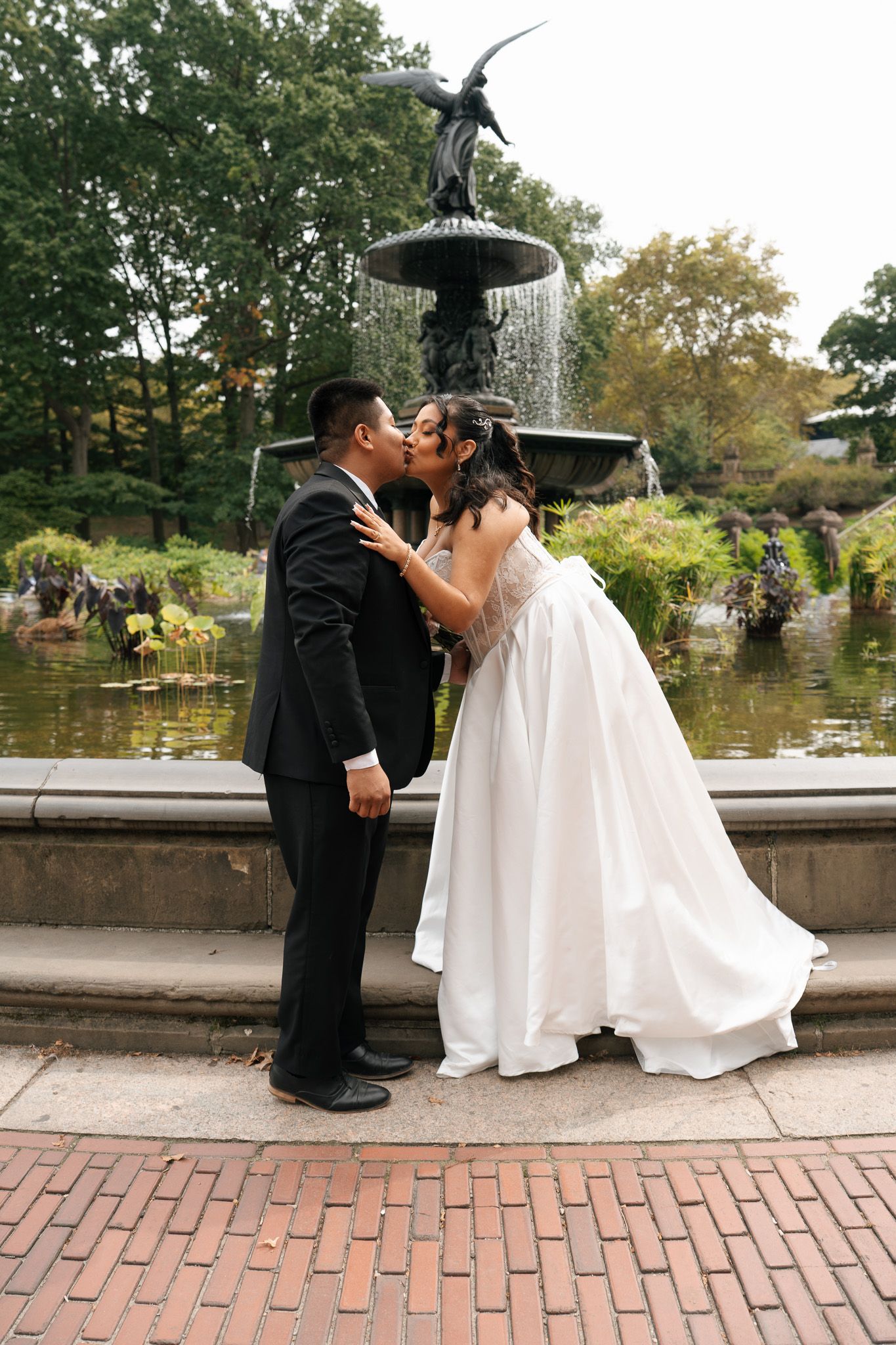 A romantic wedding couple shares a tender kiss while seated on stone steps in an outdoor garden setting.