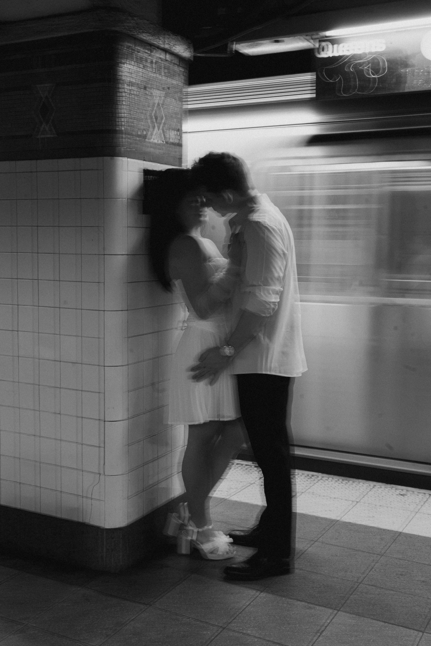 Couple sharing a romantic kiss against a tiled subway station pillar as a train blurs past in black and white.
