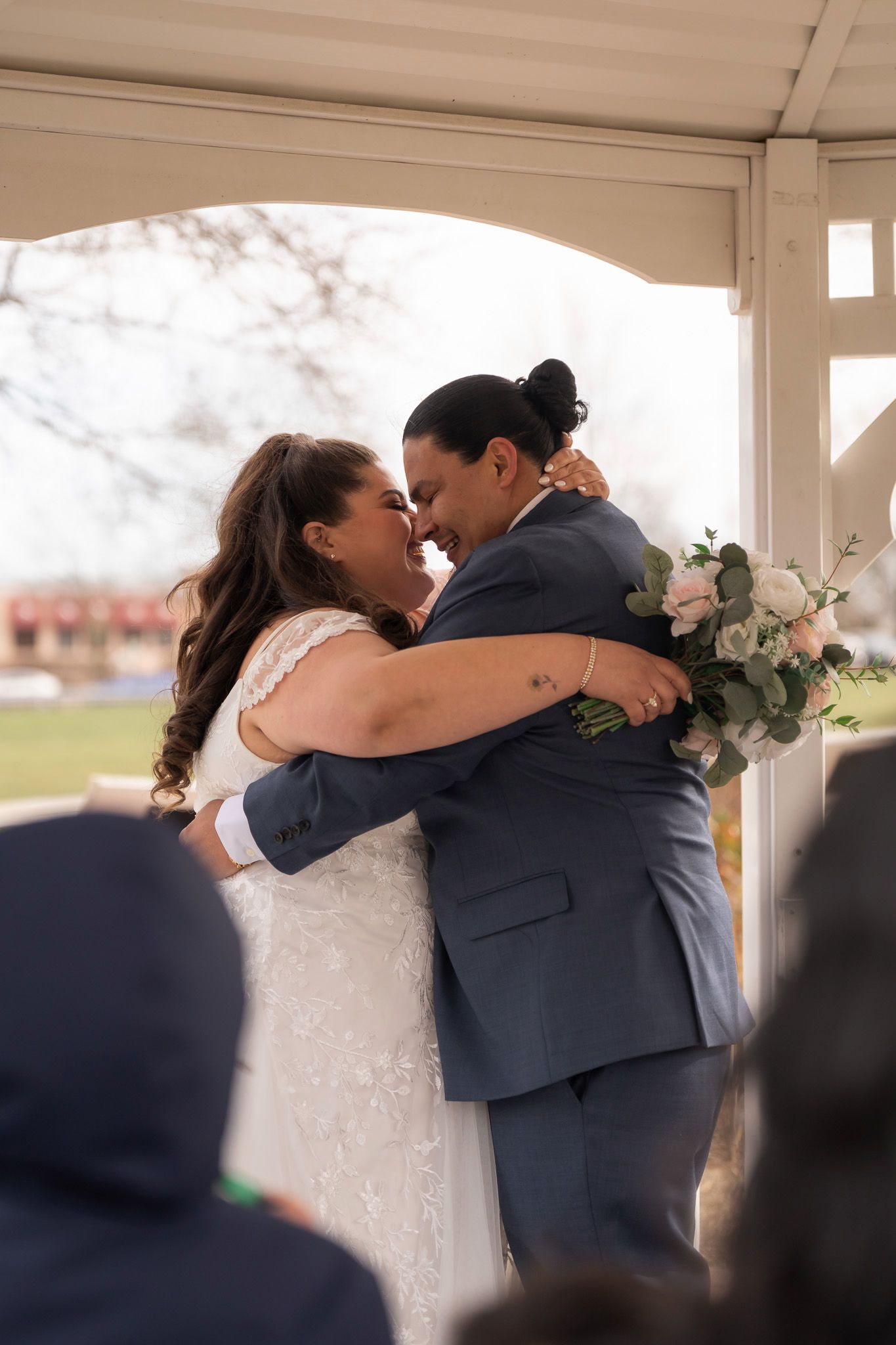 Two brides share a joyful embrace during their outdoor wedding ceremony, one in a white gown and the other in a blue suit.