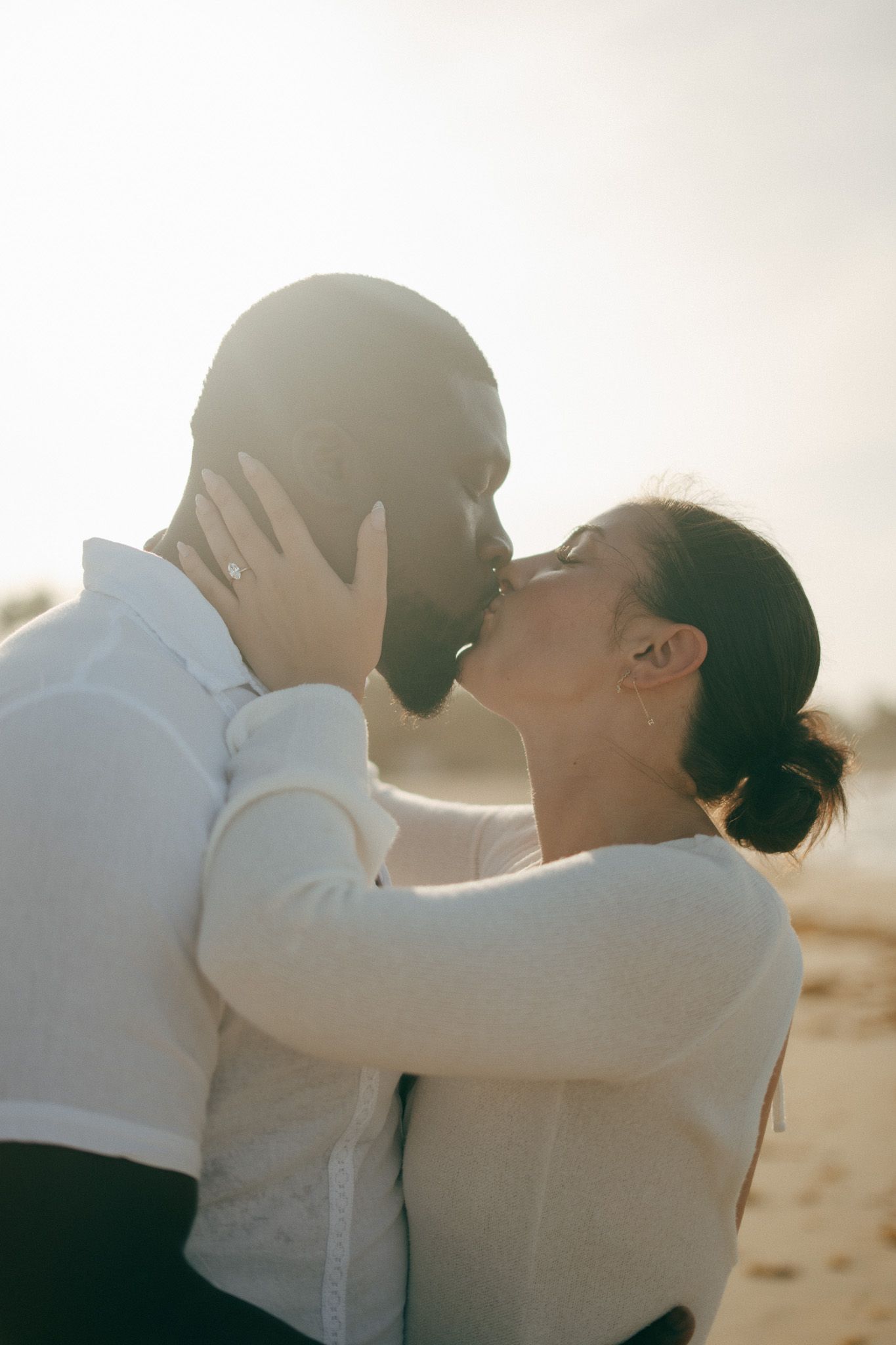 Romantic interracial couple sharing a tender kiss outdoors, backlit by warm golden sunlight.