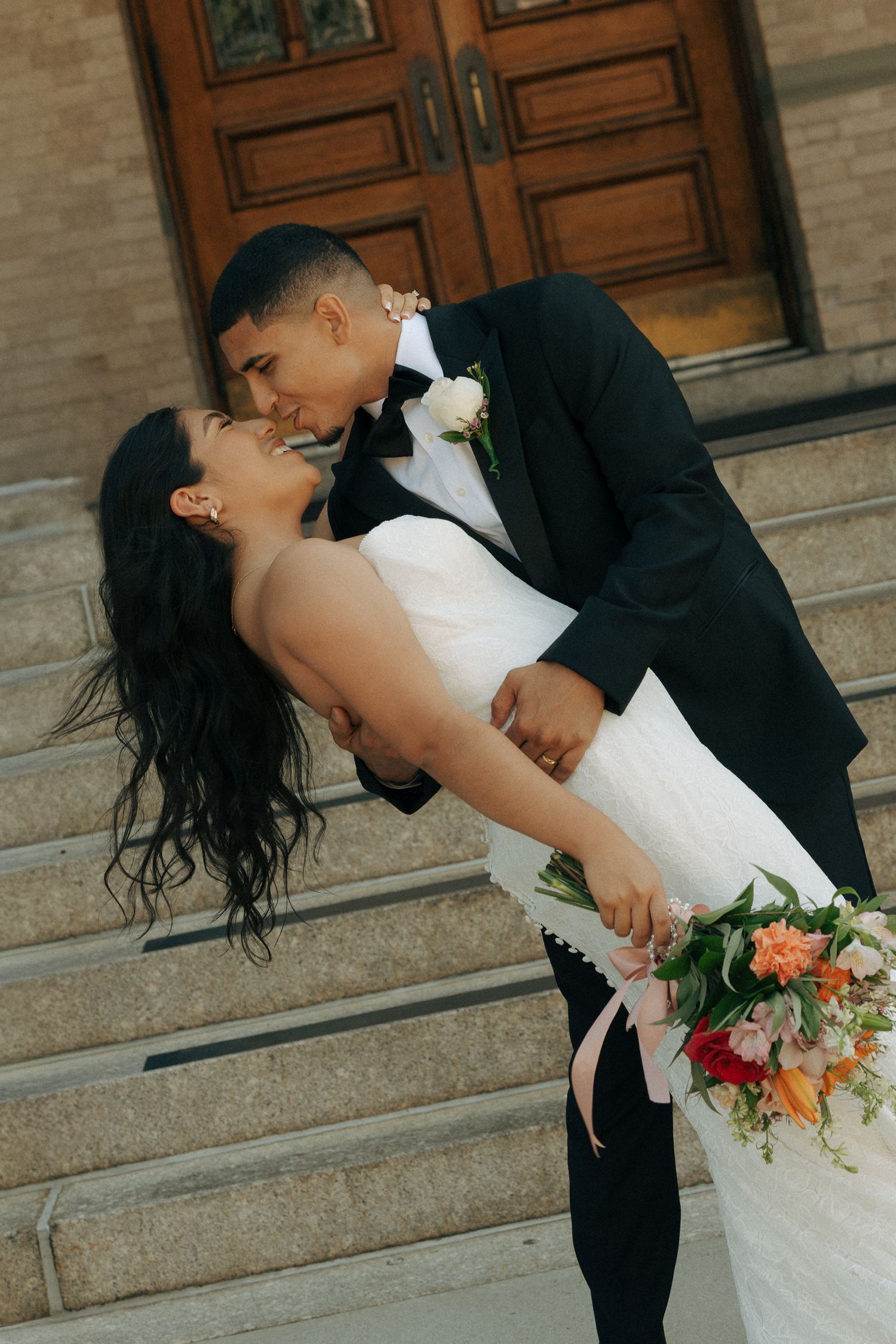 Groom dips bride in white dress holding colorful bouquet on church steps for romantic wedding kiss photo.