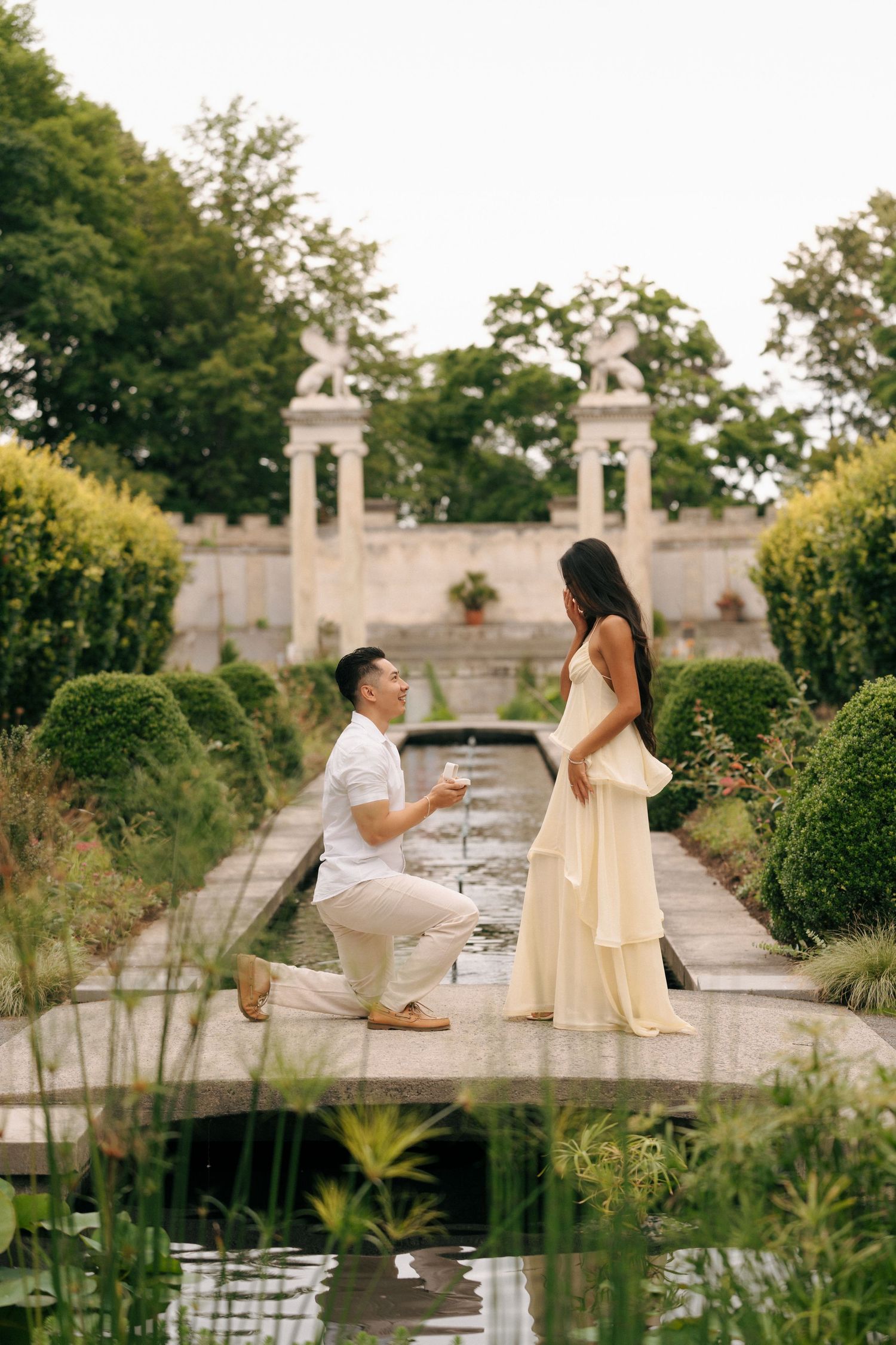 Man proposes on one knee to woman in yellow dress beside garden fountain with lush greenery and stone pillars.
