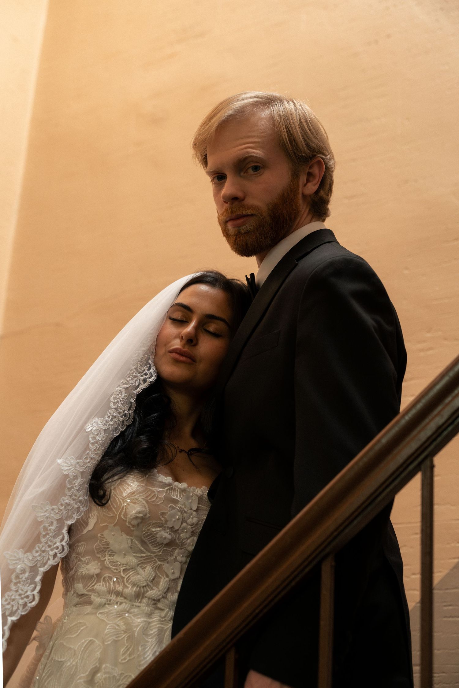 A romantic wedding couple poses on stairs in warm lighting, with the bride in white lace and groom in dark attire.