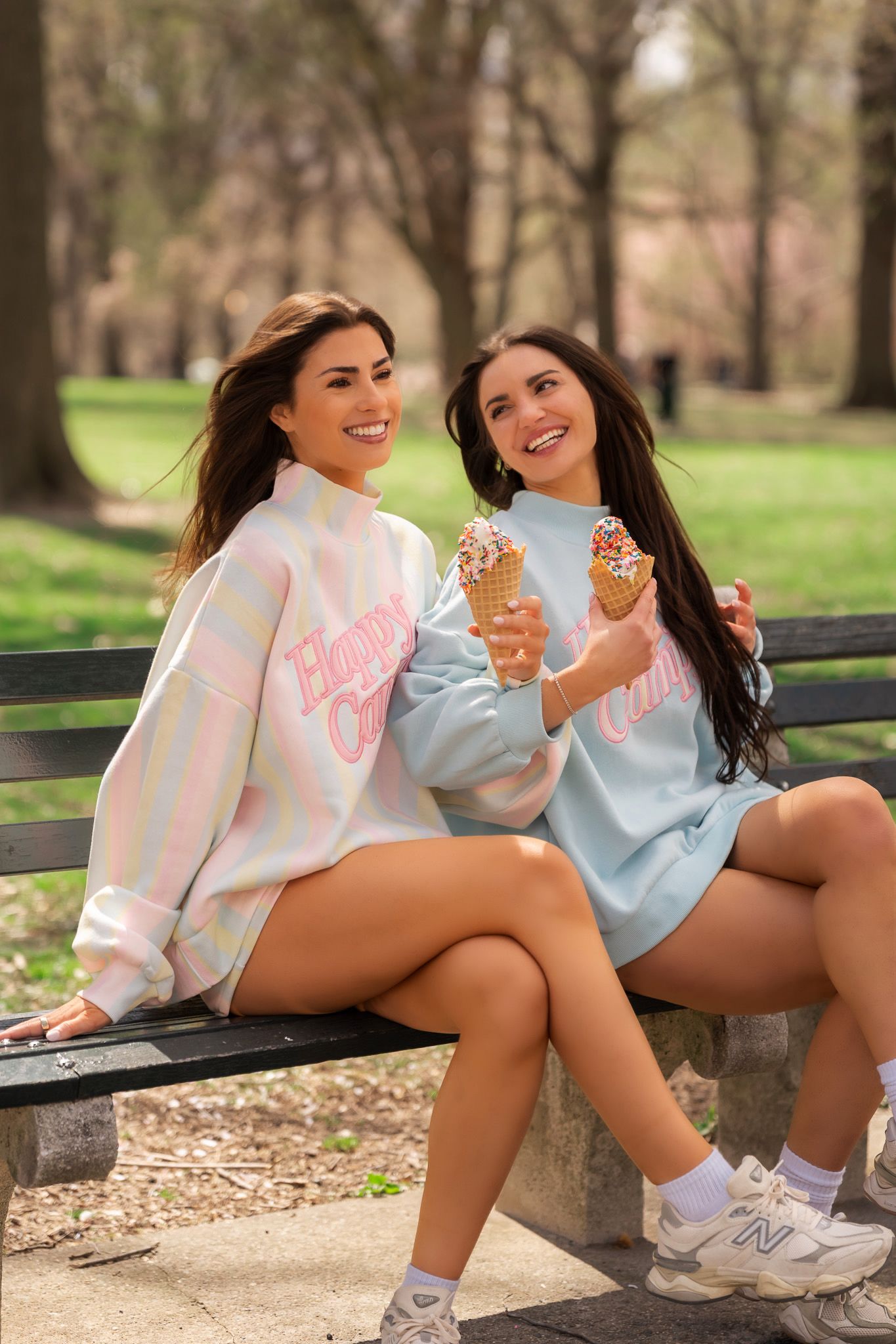 Two smiling women in pastel sweatshirts eating ice cream cones on a park bench outdoors.