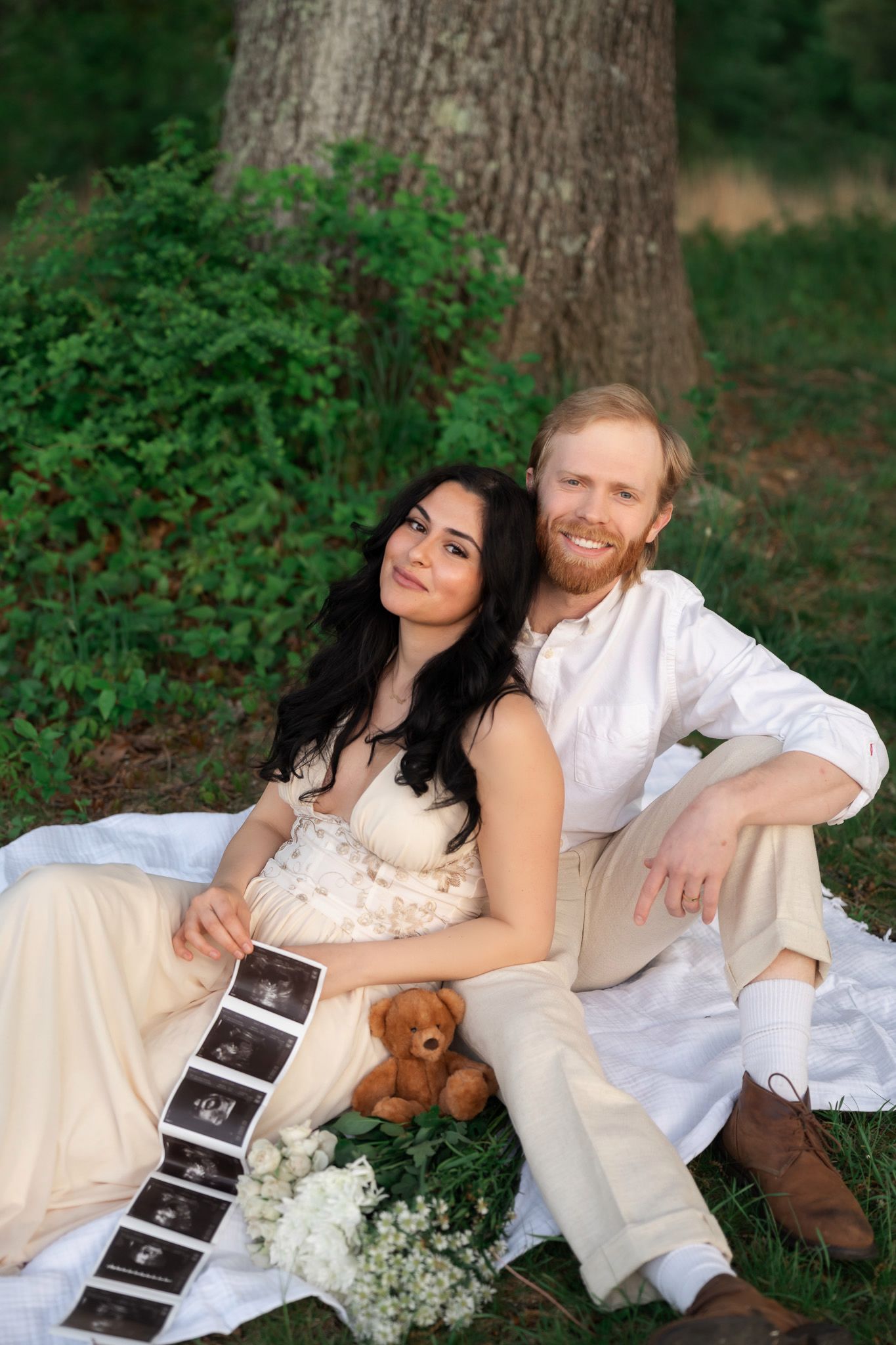 Pregnant couple sitting outdoors on blanket holding ultrasound photos with teddy bear and white flowers.