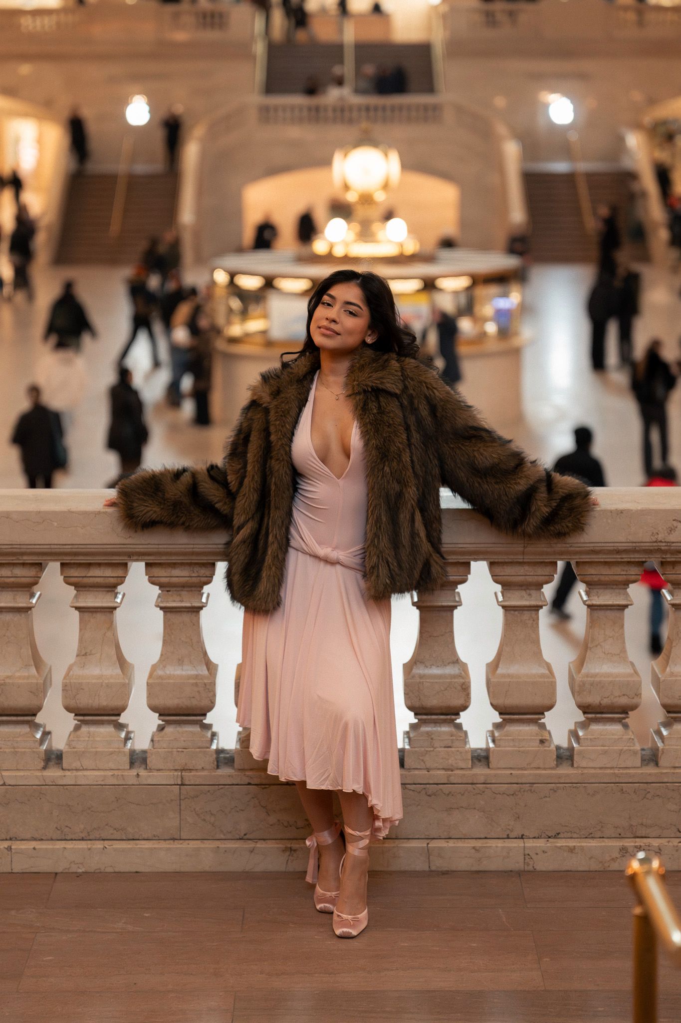 A person in a pink dress and brown fur coat poses elegantly on white marble steps at dusk.